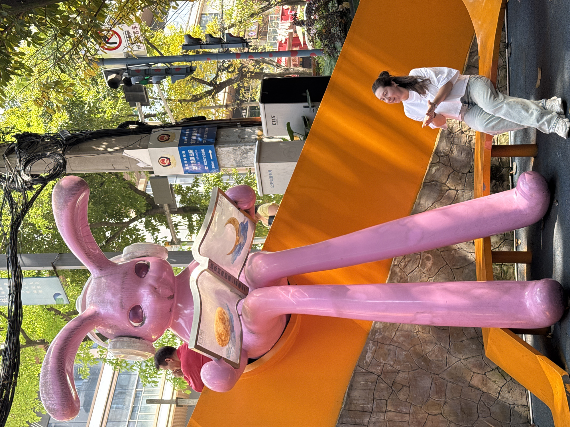 This image depicts a vibrant outdoor urban scene in Nan'an District, Chongqing, China, during bright daylight. The main subjects are a large pink rabbit sculpture and a woman interacting with it.
The prominent feature is a tall, slender pink rabbit sculpture, standing upright with long legs and arms. It wears what appear to be large, off-white headphones or earmuffs and is holding an open book with illustrative images, one resembling a crescent moon and another a golden egg or fruit. The rabbit is positioned on an orange, curved structure, possibly part of a public art installation or playground seating.
A woman, wearing a white t-shirt, light-colored jeans, and white sneakers, stands leaning against the orange structure, looking down towards the rabbit sculpture. Her hair is tied back, and she appears calm and engaged. Partially visible behind the rabbit sculpture is another person in a red shirt.
The background shows green foliage from trees, suggesting a park-like environment. A utility pole stands on the left, tangled with numerous cables. Attached to the pole are traffic lights and several signs. One sign, white with a red circle and diagonal line, indicates "no parking" or "no stopping," with the character "车" (car) visible. Another blue sign clearly states "重庆市公安局" (Chongqing Public Security Bureau) and "龙门浩派出所" (Longmenhao Police Station), along with "POLICE STATION" in English, a phone number "62872396," and "300米" (300 meters), indicating the distance to the police station. Below this, a grey utility box is labeled "公安交通专用" (Public Security Traffic Use) and "ETCS." The ground is paved with patterned stone. The scene suggests a public space, possibly a pedestrian area or park, adorned with playful art.