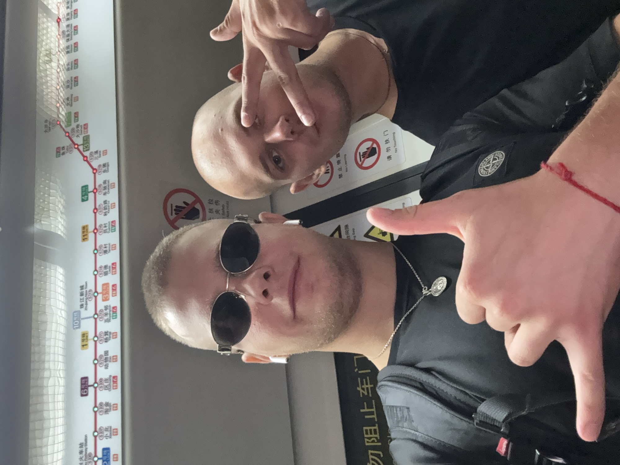 Two young bald Caucasian males are taking a selfie in a subway car, likely in Panyu District, China, given the visible Panyu District subway map in the background. The male in the foreground on the left is wearing sunglasses, a black t-shirt, a silver chain with a medallion, and a black backpack. He is giving a thumbs-up gesture with his right hand, which has a red string bracelet on his wrist. The male on the right is also wearing a black t-shirt and making a peace sign with his right hand. Both individuals appear to be in their early to mid-twenties. The subway car interior is visible, with a map displaying subway lines and station names in Chinese and English. Safety signs with hand symbols indicating "No Touching" and "No Leaning" are also present. The lighting suggests an indoor, artificially lit environment.