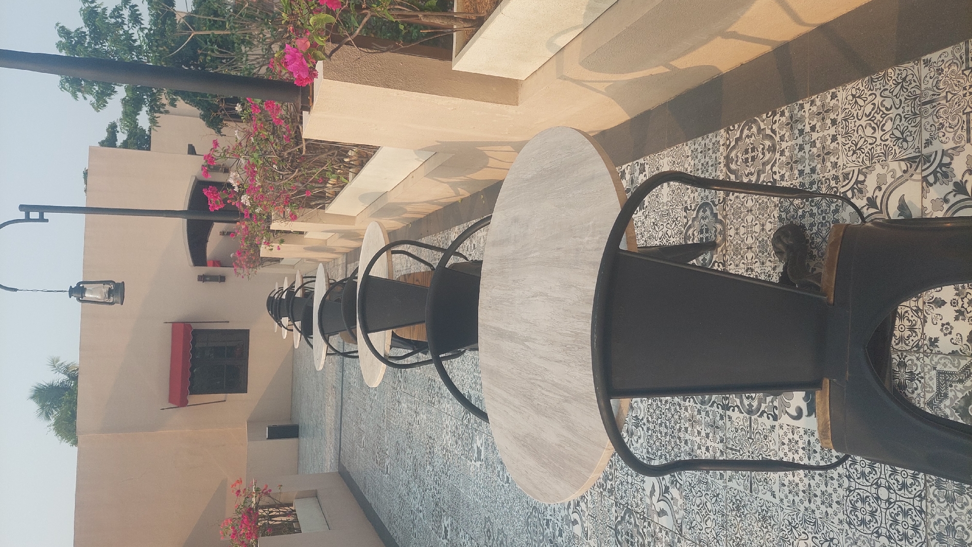 This image depicts an outdoor seating area of a cafe or restaurant in Cileungsi, Indonesia. The scene is bathed in soft, natural light, suggesting either early morning or late afternoon. The focus is on a row of empty tables and chairs, arranged in a line along a patterned tiled floor.
The tables are round with light-colored, possibly wooden or marble-effect tops and dark metal bases. They are paired with matching dark metal chairs, characterized by their simple, industrial design. The chairs have curved backrests and legs.
The setting appears to be an elevated patio or terrace, with a light-colored building wall forming the background. A small window with a red awning is visible on this wall, as well as a vintage-style lantern hanging from a black pole. Further down the row of tables, planters with vibrant pink and white bougainvillea flowers add a splash of color.
The overall atmosphere is serene and unoccupied, hinting at a quiet moment before or after peak operating hours. The tiled floor has an intricate black and white pattern, adding a decorative element to the ground. A long shadow is cast by an unseen structure or object to the right of the frame.