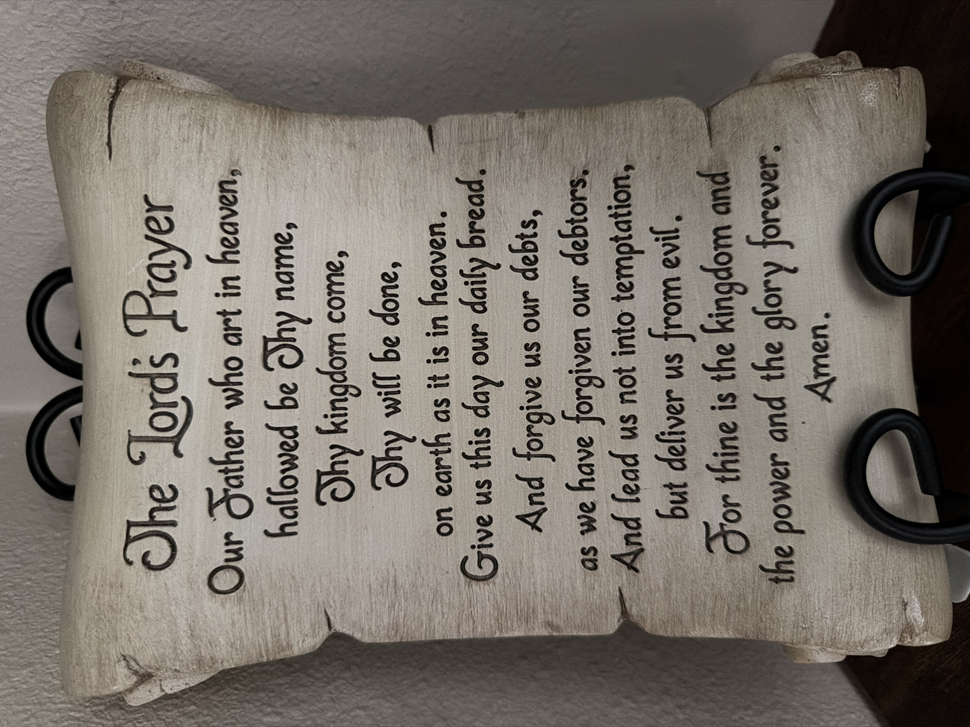 A beige, scroll-shaped plaque with the Lord's Prayer inscribed in dark lettering hangs on a light-colored wall. The plaque has distressed, aged edges and is supported by two black metal hooks at the top. The text on the plaque is in a formal script. The image is a close-up, full-frame shot of the plaque. The location in Wellford, United States of America, is not determinable from the image itself.