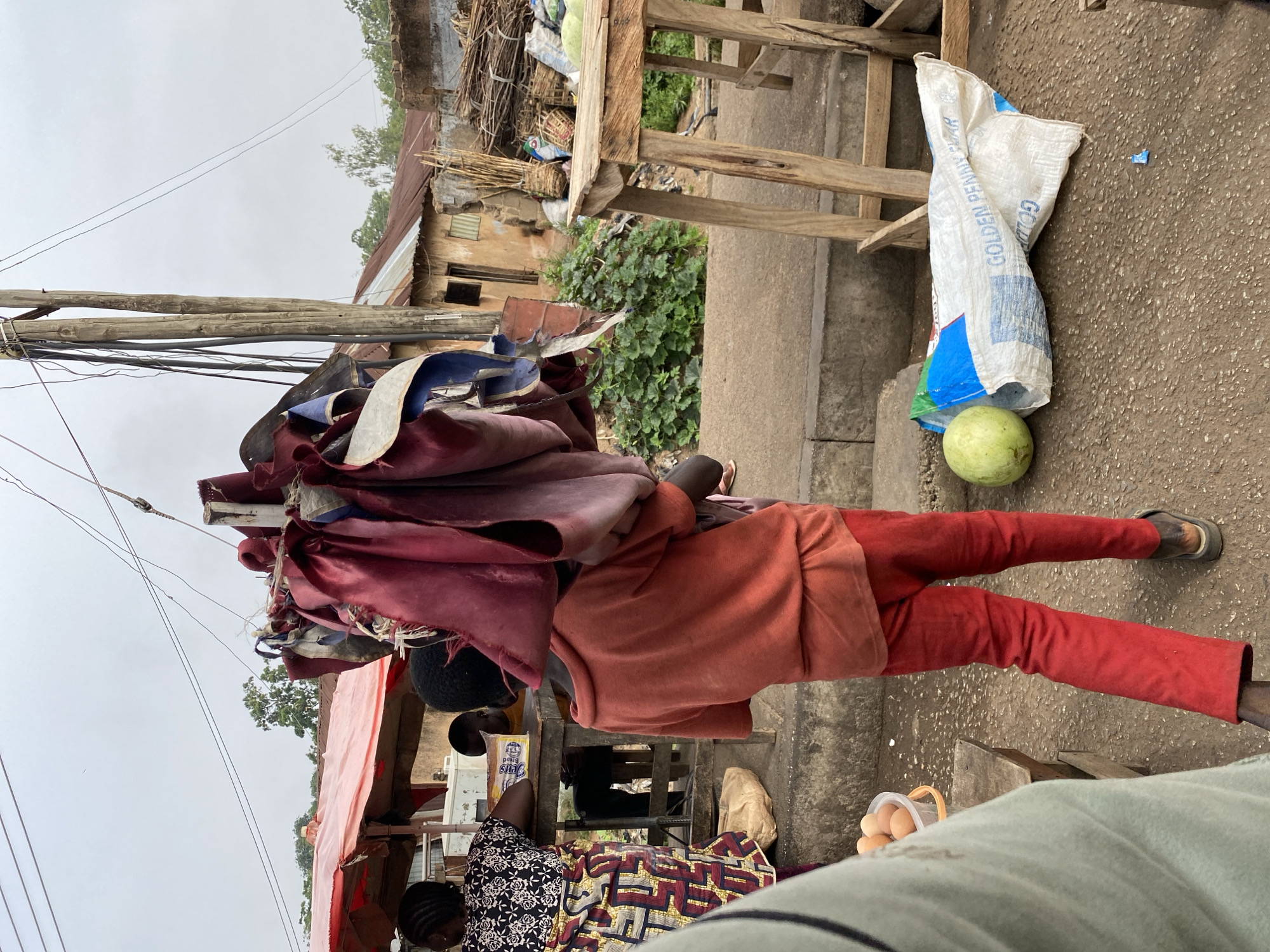The image depicts a street scene in Bukuru, Nigeria. An individual wearing red pants and an orange shirt carries a large load of fabric or material on their head. The person is walking on a concrete surface near a wooden table and a white sack labeled "GOLDEN PENALTY YEAR". Beside the sack, there's a green fruit, potentially a melon. In the foreground, there's a container of eggs and a portion of a person's clothing. A woman in a patterned dress is visible near a stall with various items. The background shows buildings, vegetation, and utility poles with wires. The sky appears overcast.