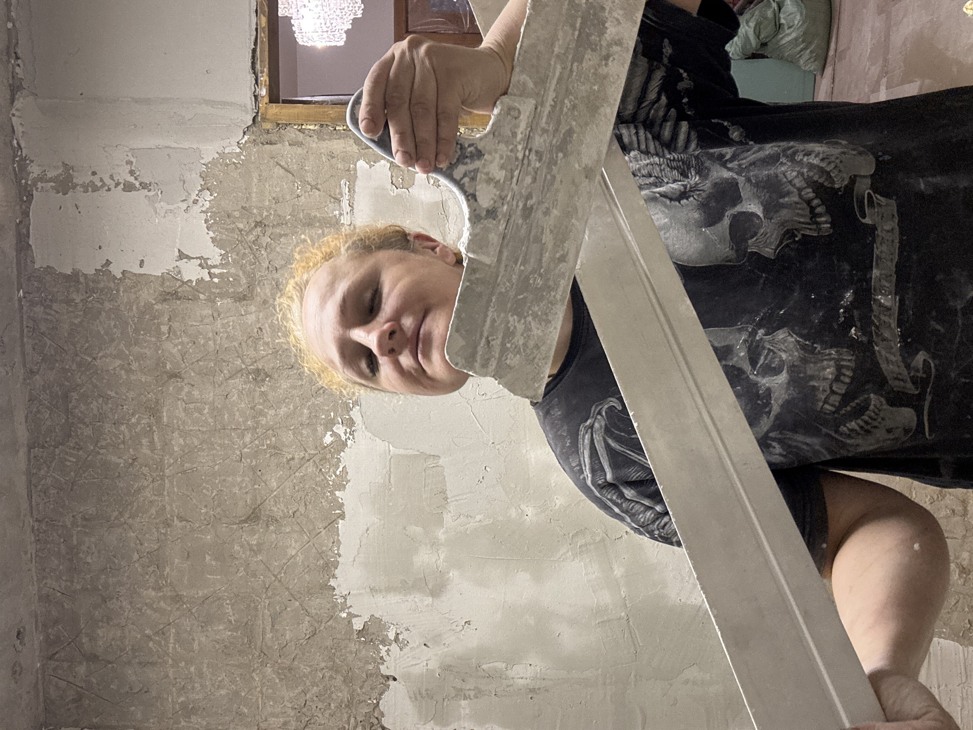 An eye-level shot captures a person engaged in wall renovation work indoors. The subject, seen from the chest up and turned partially to their right, has light curly hair and a focused, perhaps slightly weary, expression, looking downwards. They are wearing a dark t-shirt featuring a prominent white graphic of a skull and skeleton on the front, with an arm also visible adorned with a similar skeletal design.
The person is holding a large, rectangular plastering trowel or float, which is covered in dried plaster or spackle, indicating active use. The tool extends across the frame, partially obscuring the lower half of the person's face and body. The walls in the background are in a state of disrepair, with large patches of paint or plaster peeling off, revealing a rough, textured underlying surface. Some areas of the wall appear freshly spackled or plastered in a lighter tone, contrasting with the darker, exposed sections.
In the upper background, a portion of a window frame with a golden or light brown trim is visible, and through it, a sparkling chandelier can be seen, suggesting an adjacent room or a light fixture within the same space. The overall setting is an interior room undergoing construction or renovation. No specific time of day or weather conditions are discernible, and there is no visible text. The person's posture and the tool suggest they are actively working on preparing the walls.