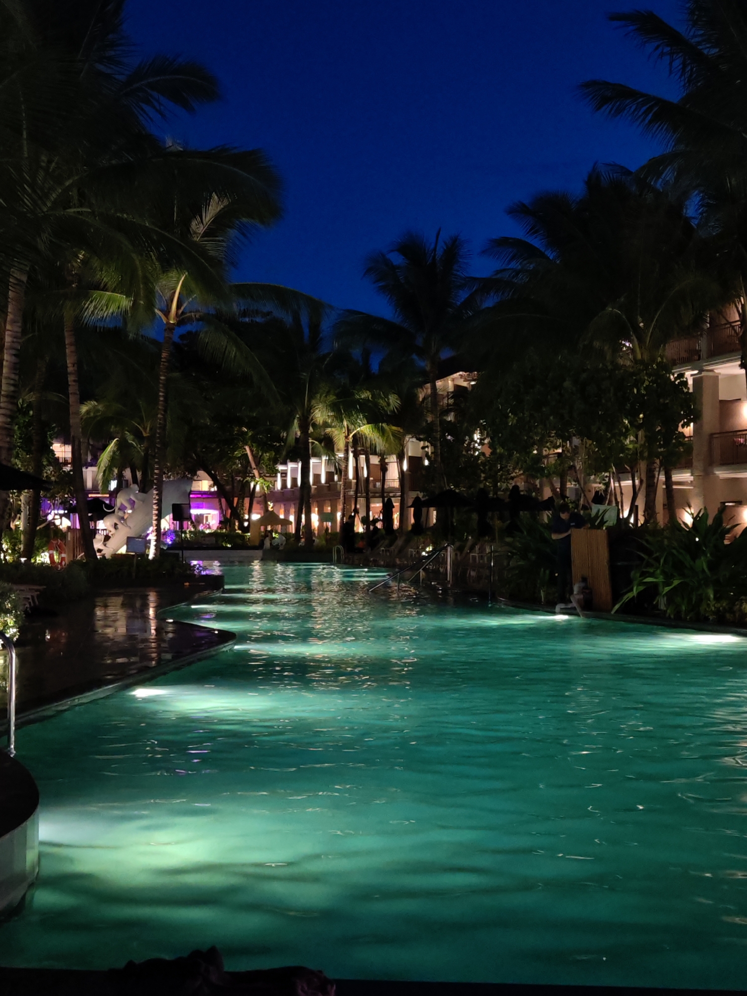 A nighttime scene depicts a swimming pool with turquoise water illuminated by underwater lights. Palm trees with dark green fronds are silhouetted against a deep blue sky, their trunks visible. In the background, a multi-story building with balconies and lit windows suggests a resort or hotel. One person is visible in the pool area, near a cluster of plants. The area surrounding the pool appears to be a patio or deck. The image is taken in Baan Bo Phut, Thailand.