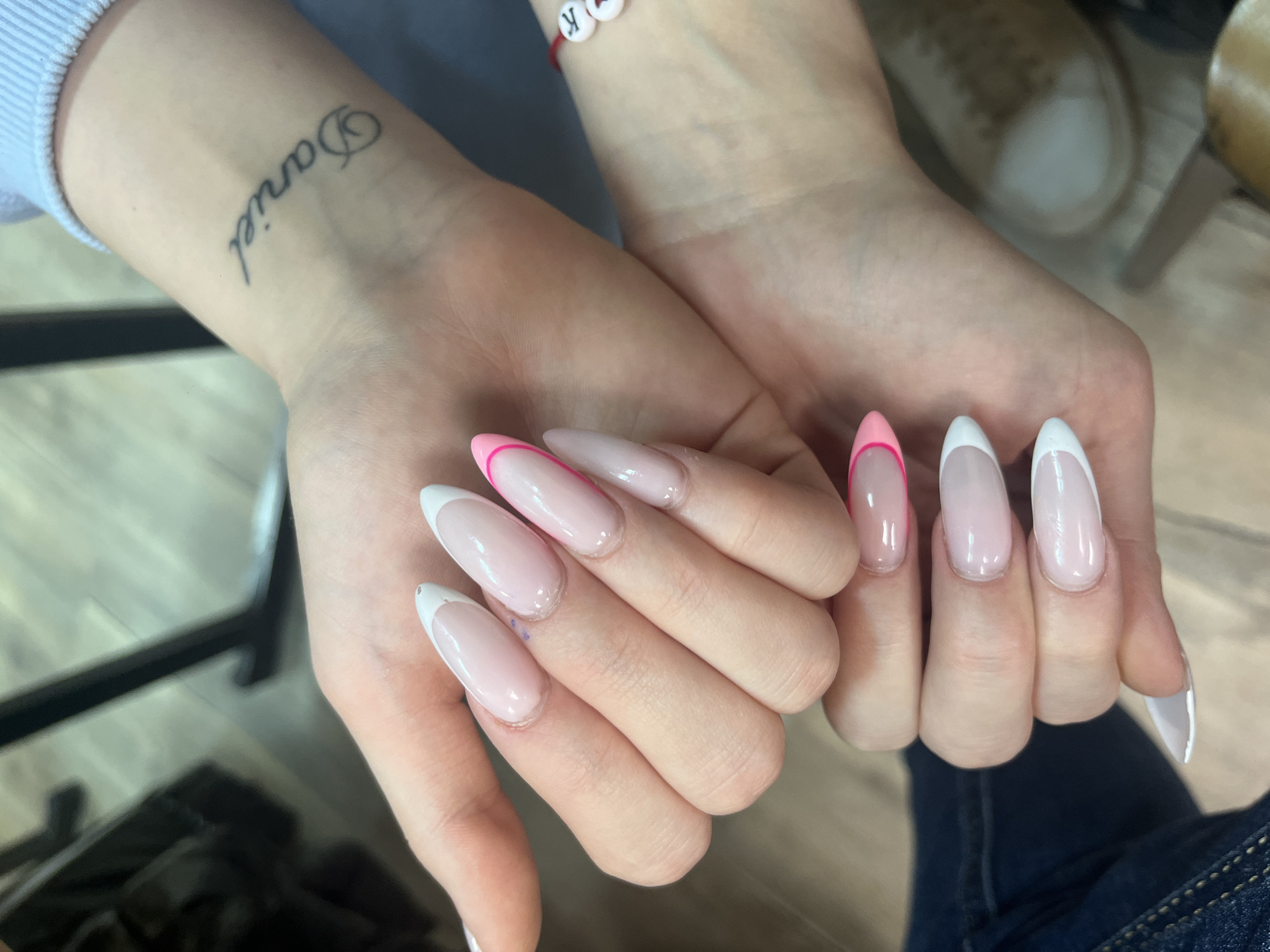 The image shows a person's hands with manicured nails. The nails are long and almond-shaped, with a nude or pale pink base color. Some nails have a classic white French tip, while others have a bright pink tip. The person has a tattoo on their left wrist that appears to read "Daniel" in cursive. A red string bracelet with white beads bearing the letters "K" and "R" is visible on the same wrist.
The background is blurred, but some details are discernable. There are hints of furniture, possibly a chair or table, and clothing. A shoe is visible on the top right, giving more clues about the setting. The location is not confirmed.