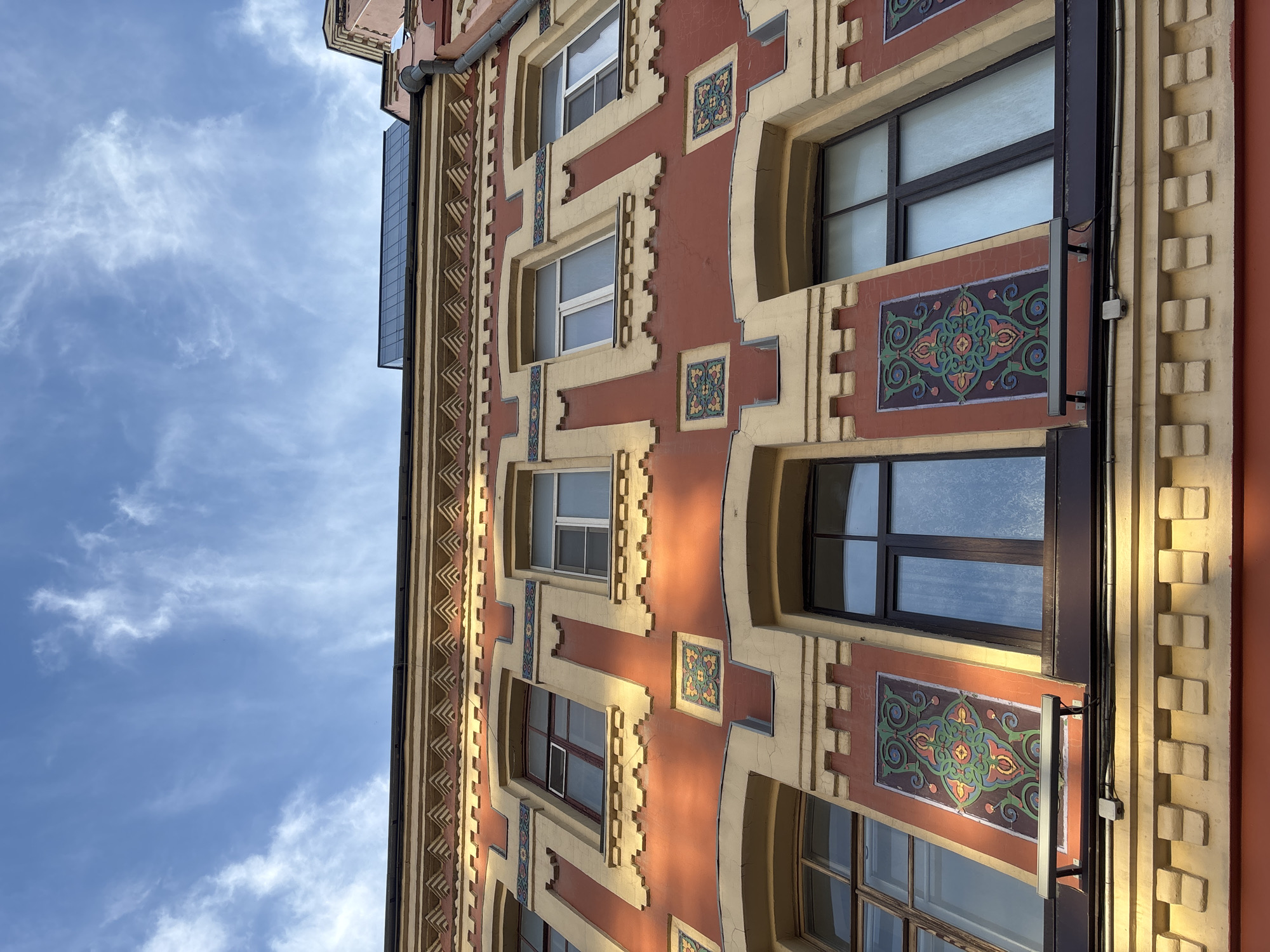 This is a low-angle, exterior, eye-level shot of an ornate building facade. The building has a red base color with yellow decorative trim. The trim features geometric patterns, and there are square decorative tiles with intricate colorful patterns set into the red facade. Several windows are visible, some with dark frames and others with lighter frames, and some are multi-paned. The sky above is blue with wispy white clouds. The lighting suggests it is either early morning or late afternoon, with sunlight casting shadows on the building. The building appears to be located in Novaya Usman', Russia.