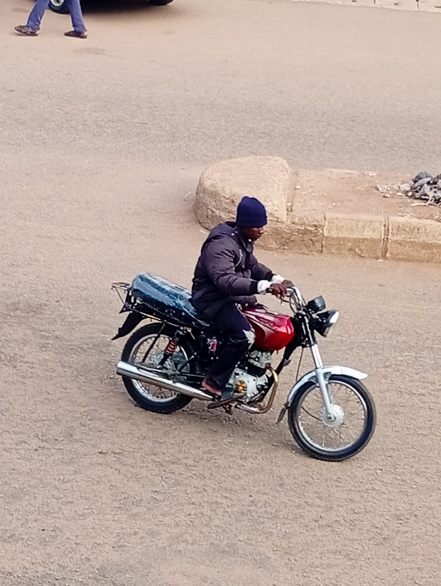A person is riding a red and black motorcycle on a dirt road in Bukuru, Nigeria. The person is wearing a blue beanie, a dark puffer jacket, and dark pants. The motorcycle has a chrome exhaust pipe, silver spokes, and a red gas tank. In the background, there is a large rock and some scattered debris. A person's legs and feet are visible in the top left corner of the image, walking on the dirt road.