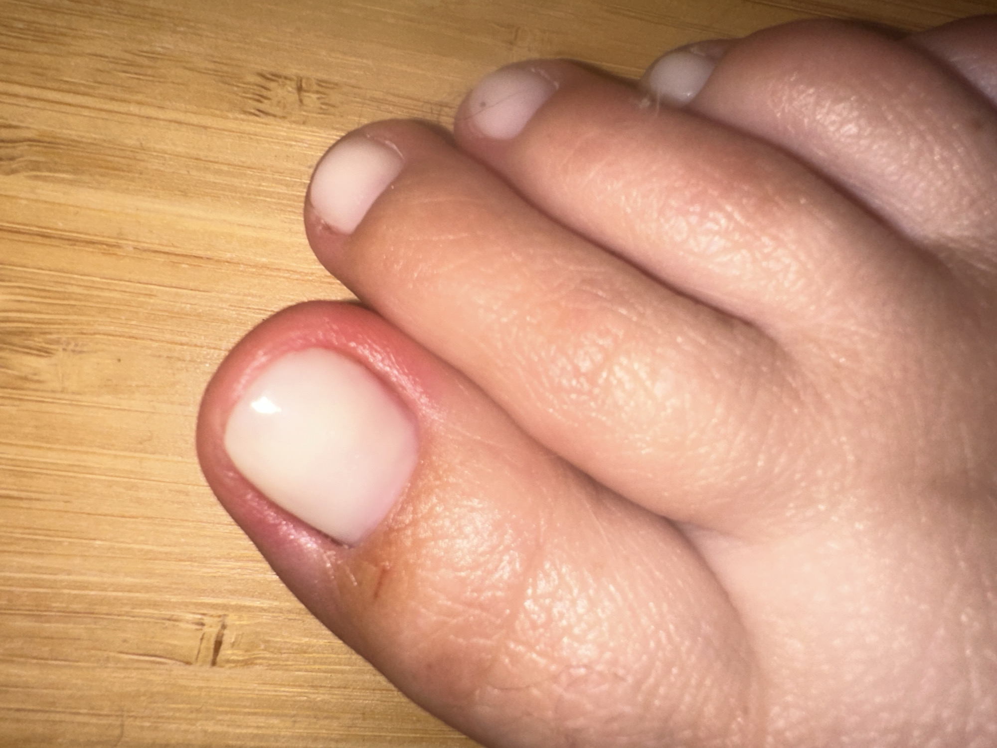A close-up, eye-level, medium shot shows a person's left foot against a wood-grained background. The foot is positioned diagonally, with the big toe pointing towards the upper left corner of the frame. The toenails appear to be freshly painted or naturally healthy and smooth, with a light, pearlescent sheen. The skin of the foot is light-skinned, with visible texture and fine lines. There is a small, dark mark, possibly a bruise or a speck of dirt, on the skin near the cuticle of the big toe. The other toes are partially visible, fanned out and resting on the wooden surface. The wood grain background is composed of vertical lines with subtle variations in color, suggesting natural bamboo or a similar material.