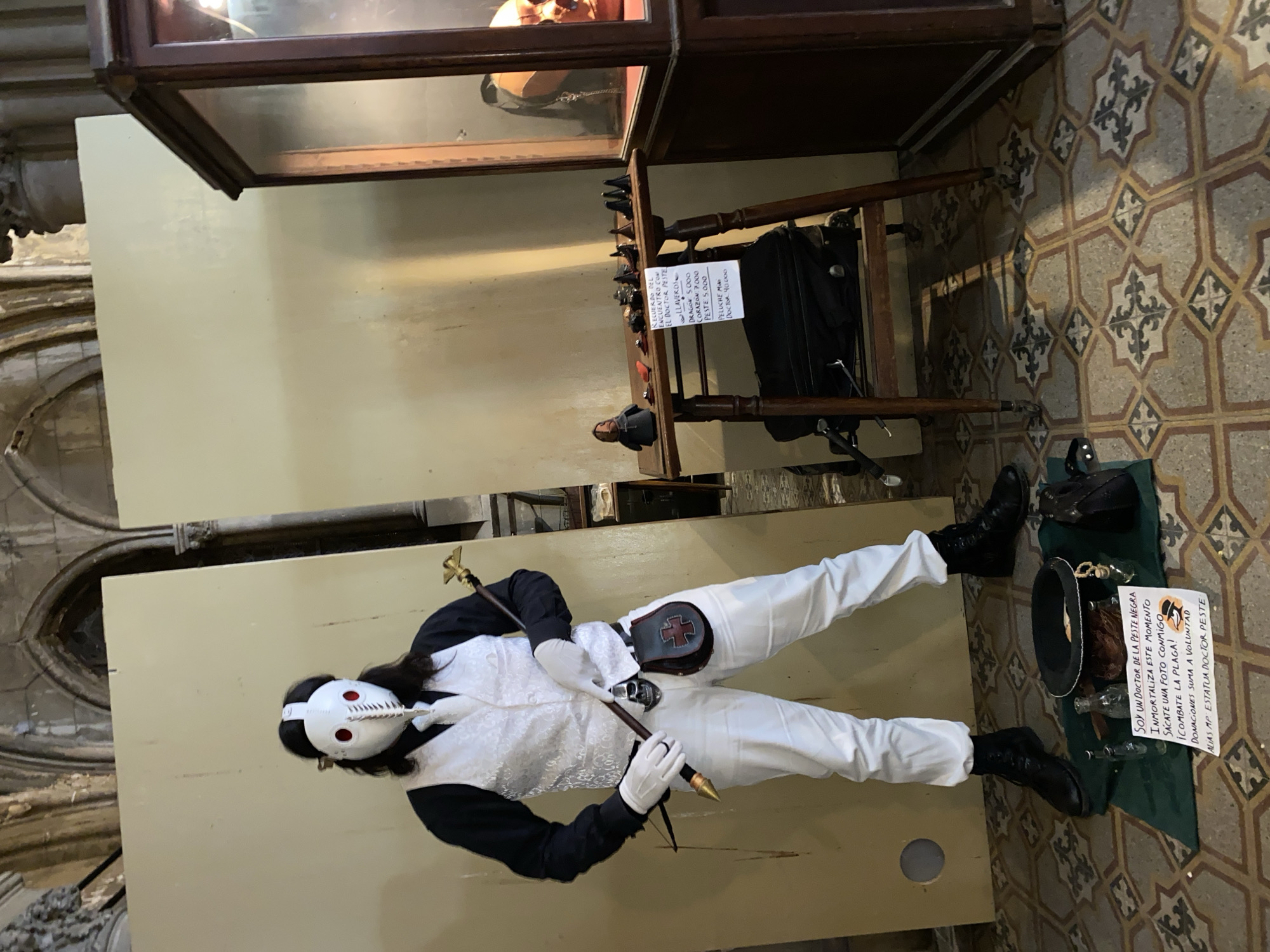 The image shows a person dressed as a plague doctor, lying on a table in what appears to be a museum or historical building, located in Buenos Aires, Argentina, based on the Spanish text. The plague doctor is wearing a white mask with red-rimmed eye holes, a black jacket with white vest, white pants, and black boots. They are holding a cane. Next to them is a green cloth with a black hat, glass bottles, and a sign written in Spanish that translates to "I am a doctor of the Black Death. Immortalize this moment. Take a photo with me. Fight the plague! Donations are welcome. Alias MP Statue Doctor Peste." Above the table are museum displays, including a glass case and a wooden stand holding items for sale, such as keychains with various shapes and prices listed. The floor is tiled with a geometric pattern.