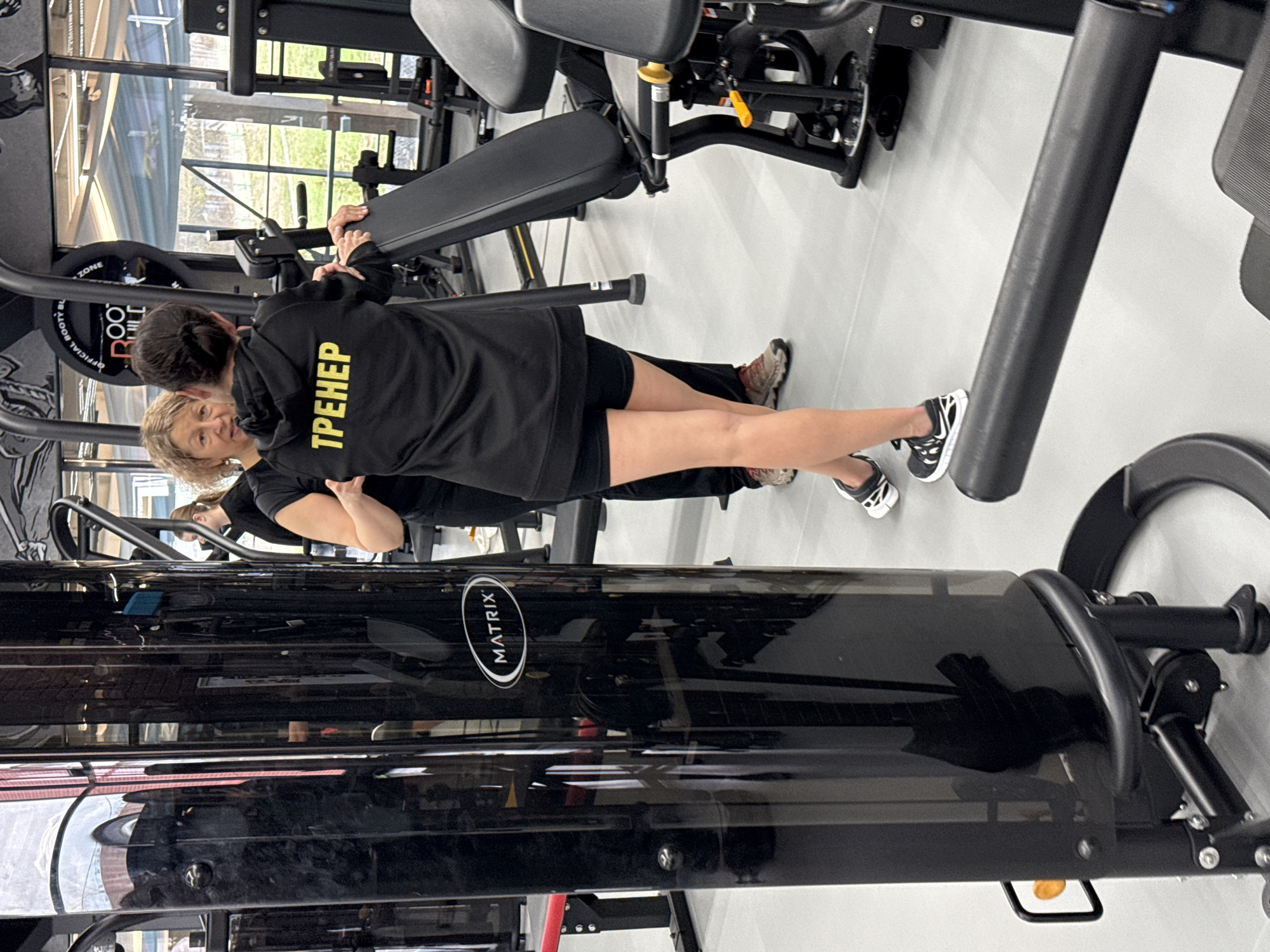 Two women are captured in an indoor gym setting in Votkinsk, Russia. One woman, with dark hair, stands with her back mostly to the camera, wearing a black long-sleeved top and black shorts. On the back of her top, the word "ТРЕНЕР" (TRAINER in Russian) is prominently displayed in yellow letters. She appears to be interacting with or assisting the second woman.
The second woman, with light, curly hair, is partially visible, facing towards the first woman with a slight smile on her face. She also wears a black top and is positioned near a black gym machine, possibly a leg press or similar equipment.
The gym features modern black exercise equipment, including a machine with a visible "MATRIX" logo. In the background, large windows reveal green foliage outside, indicating it is daytime. Another piece of equipment has "OFFICIAL BOOTY ZONE" written on a circular weight plate. The floor is a light gray or white. The overall scene suggests a personal training session or guidance in a fitness facility.
