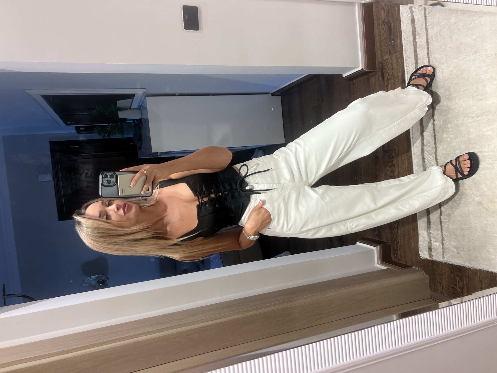In the city of Sharm El Sheikh, Egypt, a blonde woman with long hair is taking a mirror selfie in what appears to be a dressing room or bedroom. She is wearing a black lace-up corset top and high-waisted, loose-fitting white pants. Her left hand is casually tucked into her pants pocket, and she is wearing black strappy sandals. The background shows a large mirror, a wooden cabinet, a white dresser with decorative items, and part of a white closet or wall. A wooden floor is visible beneath a light-colored rug. The lighting is dim, creating a moody atmosphere.