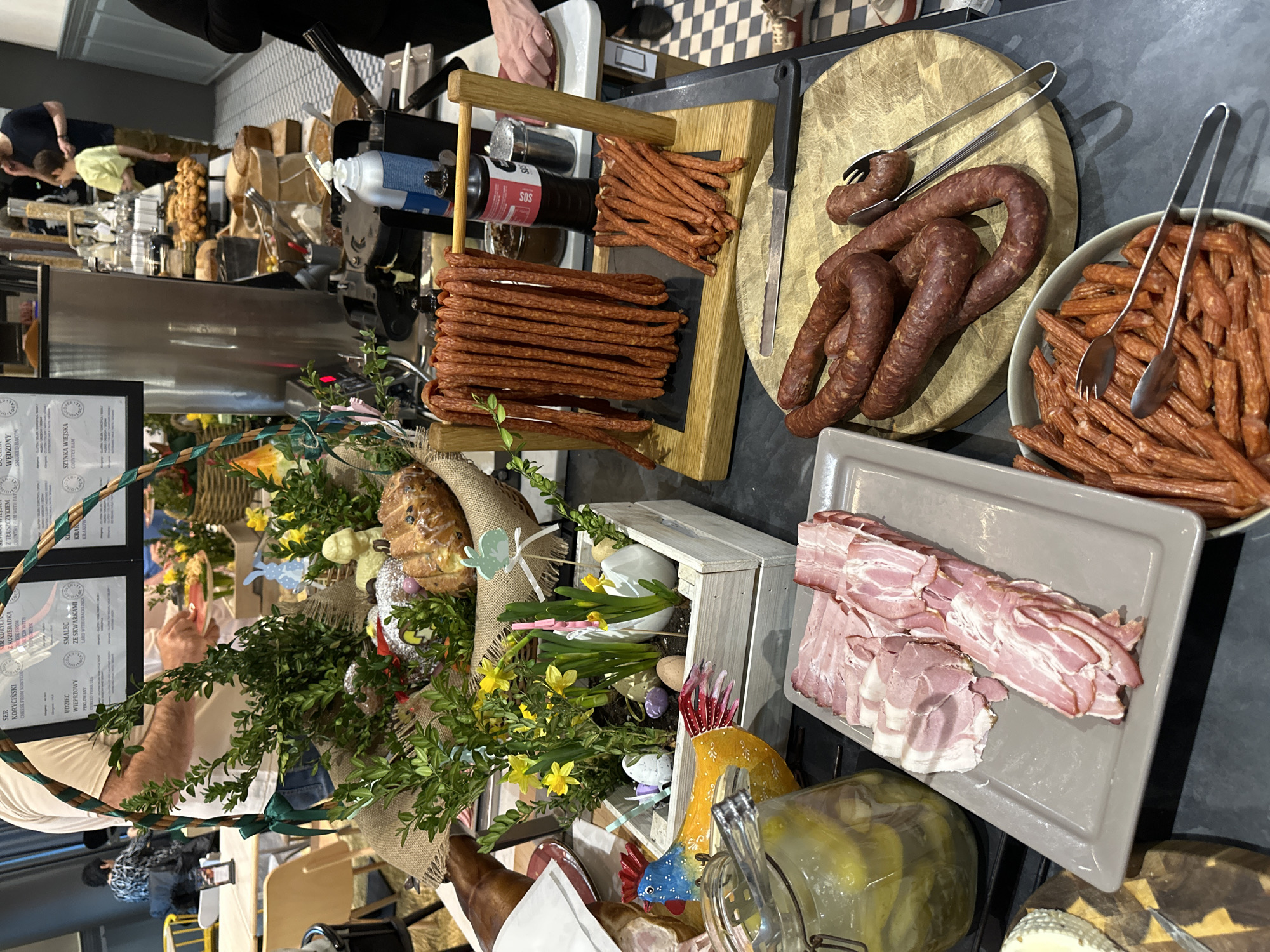 The image shows a counter display of Polish meats and Easter decorations, likely in Wrocław, Poland. There are various types of sausages, sliced bacon, a large Easter basket filled with baked goods and decorations like yellow flowers, eggs, and figurines. A wooden crate with daffodils and eggs is also present. The scene includes food preparation equipment such as knives and tongs. Behind the meat display, out of focus, is a restaurant counter with people, baked goods and equipment.