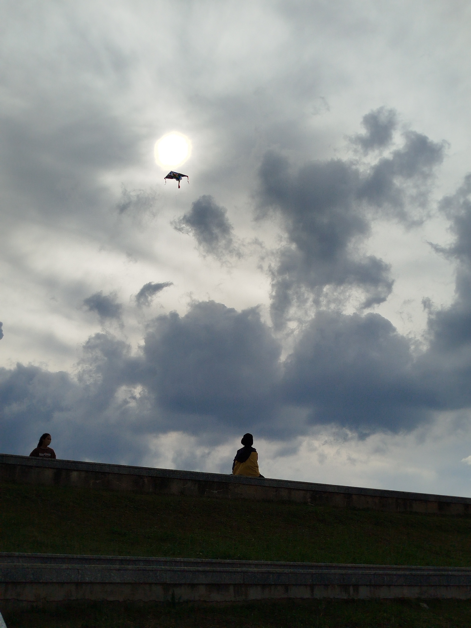 The image shows two people sitting on a wall in Da Lat, Vietnam. The sky is overcast with dark clouds and a bright sun. There is a kite flying in the sky. The people are silhouetted against the sky. The wall is made of stone and is surrounded by grass. The photo may be taken on Xuan Huong lake.