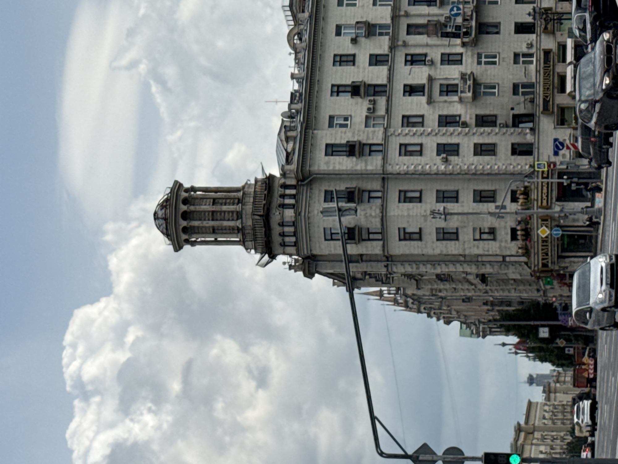 The image captures a view of the Stalinist Empire-style building on Kotelnicheskaya Embankment in Moscow, Russia, on a cloudy day. The building, characterized by its prominent corner tower topped with a spire and decorative elements, dominates the frame. Several windows, some with balconies, are visible on its facade. In the foreground, a traffic light shows a green signal, and several cars are on the street. The sky is filled with large, white cumulus clouds.