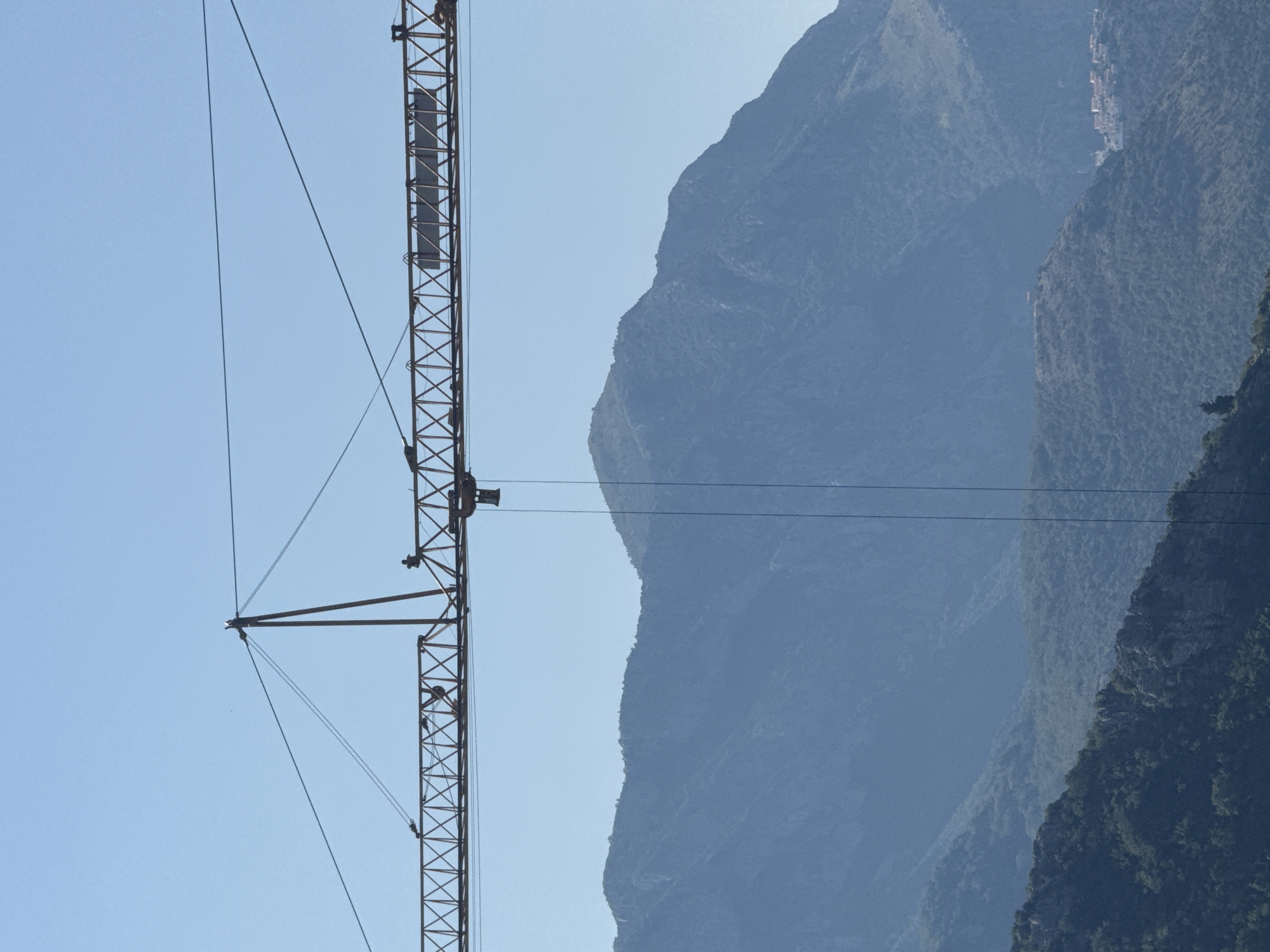 A construction crane is pictured in the foreground, its metal lattice structure extending vertically. The crane is silhouetted against a clear, pale blue sky. In the background, a series of hazy, blue-green mountains rise. The mountains are densely covered in vegetation, with their rugged slopes and peaks receding into the distance. A small cluster of buildings is visible on the side of one of the mountains, suggesting a settlement. The image appears to have been taken during the daytime, under fair weather conditions. The perspective is such that the crane dominates the left side of the frame, while the mountains fill the right. There is no visible text in the image.