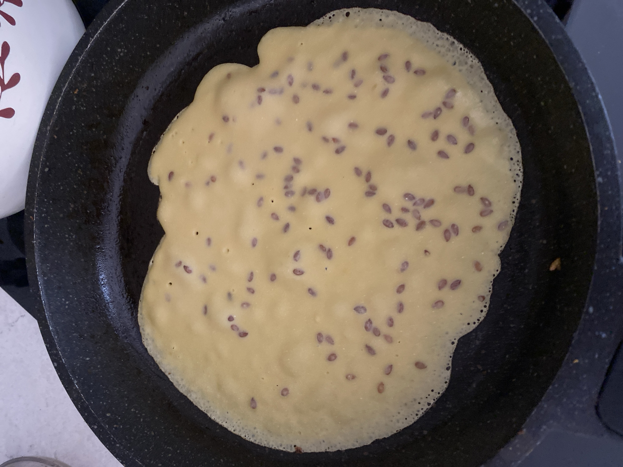 A close-up shot captures a single, thin crepe with a light yellow hue cooking in a black non-stick skillet. The crepe, irregularly shaped, is dotted with small, dark brown seeds, likely flax seeds, distributed throughout its surface. The edges of the crepe are slightly bubbly and crisping. The skillet sits on a dark surface, possibly a stovetop, with a portion of a white bowl decorated with red floral patterns visible in the upper right corner of the frame. The scene appears to be indoors, likely in a kitchen setting. There are no visible people or specific landmarks that would indicate a location in Alanya, Turkey.