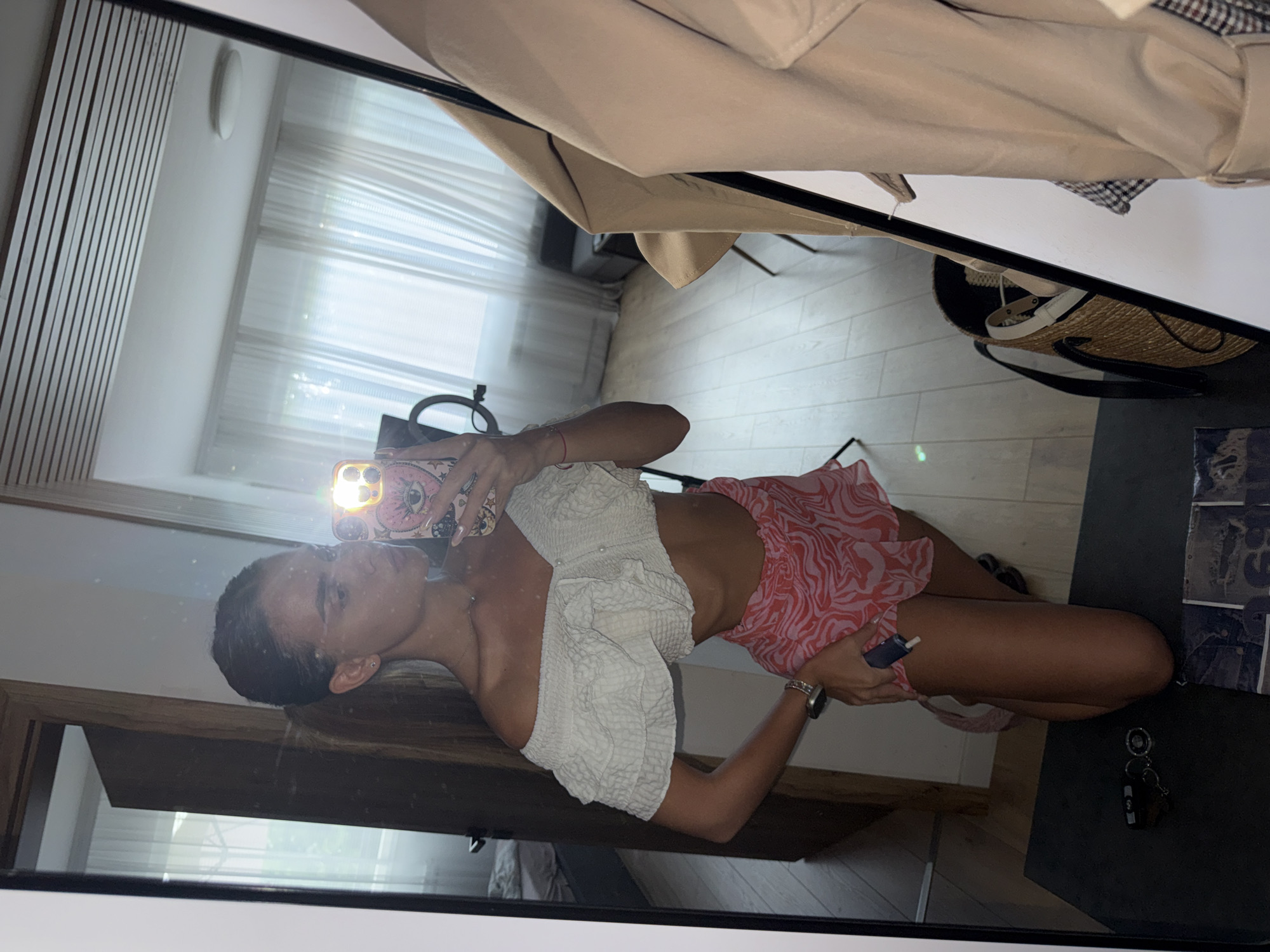 The image shows a young woman posing for a mirror selfie in what appears to be a modern apartment. She is wearing a white off-the-shoulder crop top with ruffled details and a pink and white patterned mini skirt. Her hair is pulled back in a ponytail. She is holding her phone, which has a pink case with an eye design, and her reflection is visible in a large mirror. In the reflection, we see a room with light-colored wooden floors and white curtains covering a window. There is a glimpse of a doorway to another room, a chair, and a woven straw bag on the floor. There is no indication of the city of Plovdiv, Bulgaria in the image.