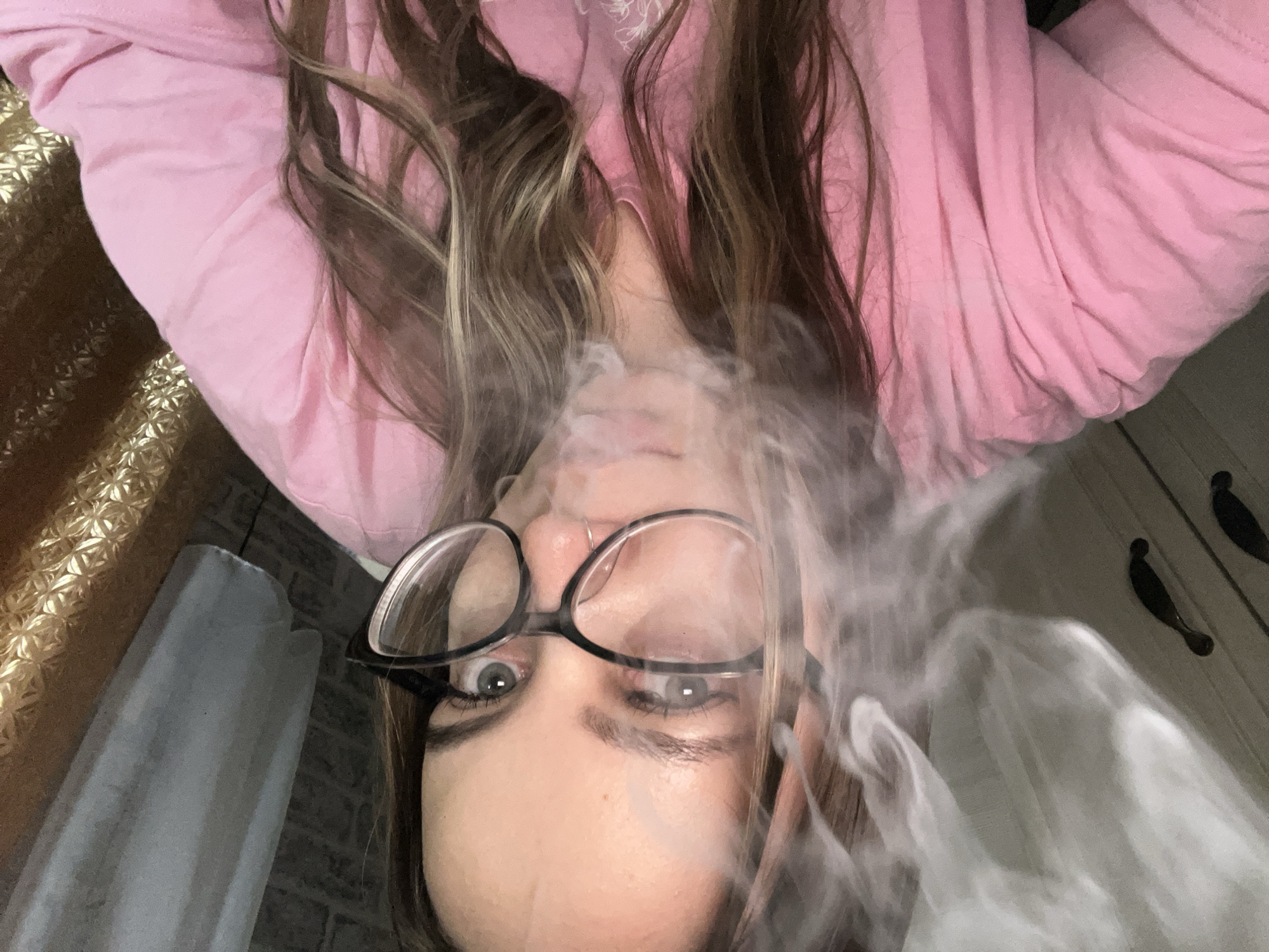 A woman with long, highlighted hair and glasses is looking directly at the camera. She is wearing a pink top. Smoke obscures part of her face. The background includes a gold-patterned surface, a gray object, and a wooden panel with dark-colored handles. The location in Novopokrovskaya, Russia, is not discernible from the image.