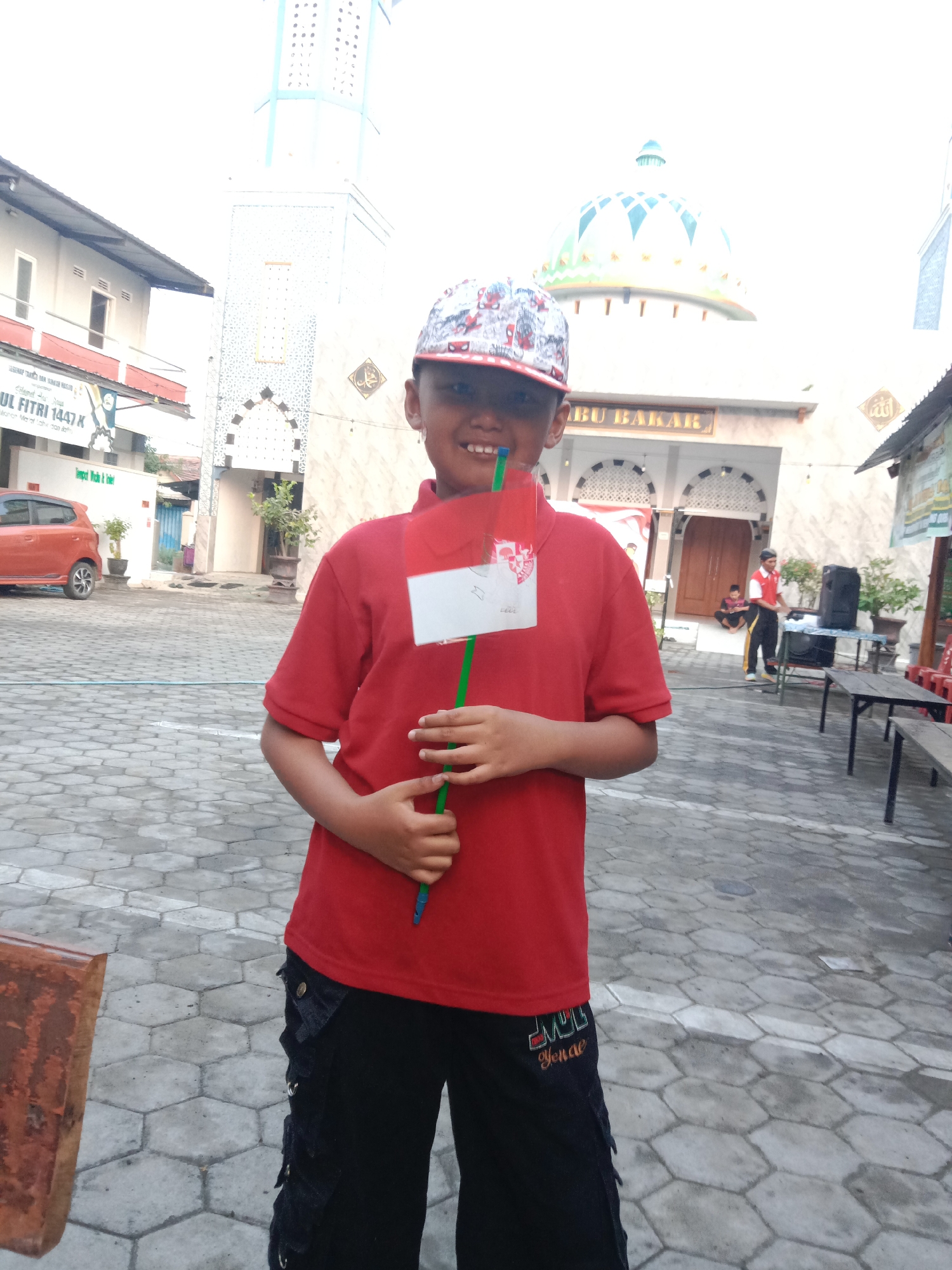 A young boy stands in front of a mosque in Madiun, Indonesia, holding a small Indonesian flag. The boy wears a red t-shirt, black pants, and a baseball cap. He is smiling and looking at the camera. The mosque has a large dome and a tall minaret. The courtyard is paved with hexagonal tiles, and there are trees and plants around. In the background, a few other people can be seen, and there are buildings and a car. The sky is bright, suggesting it is daytime.