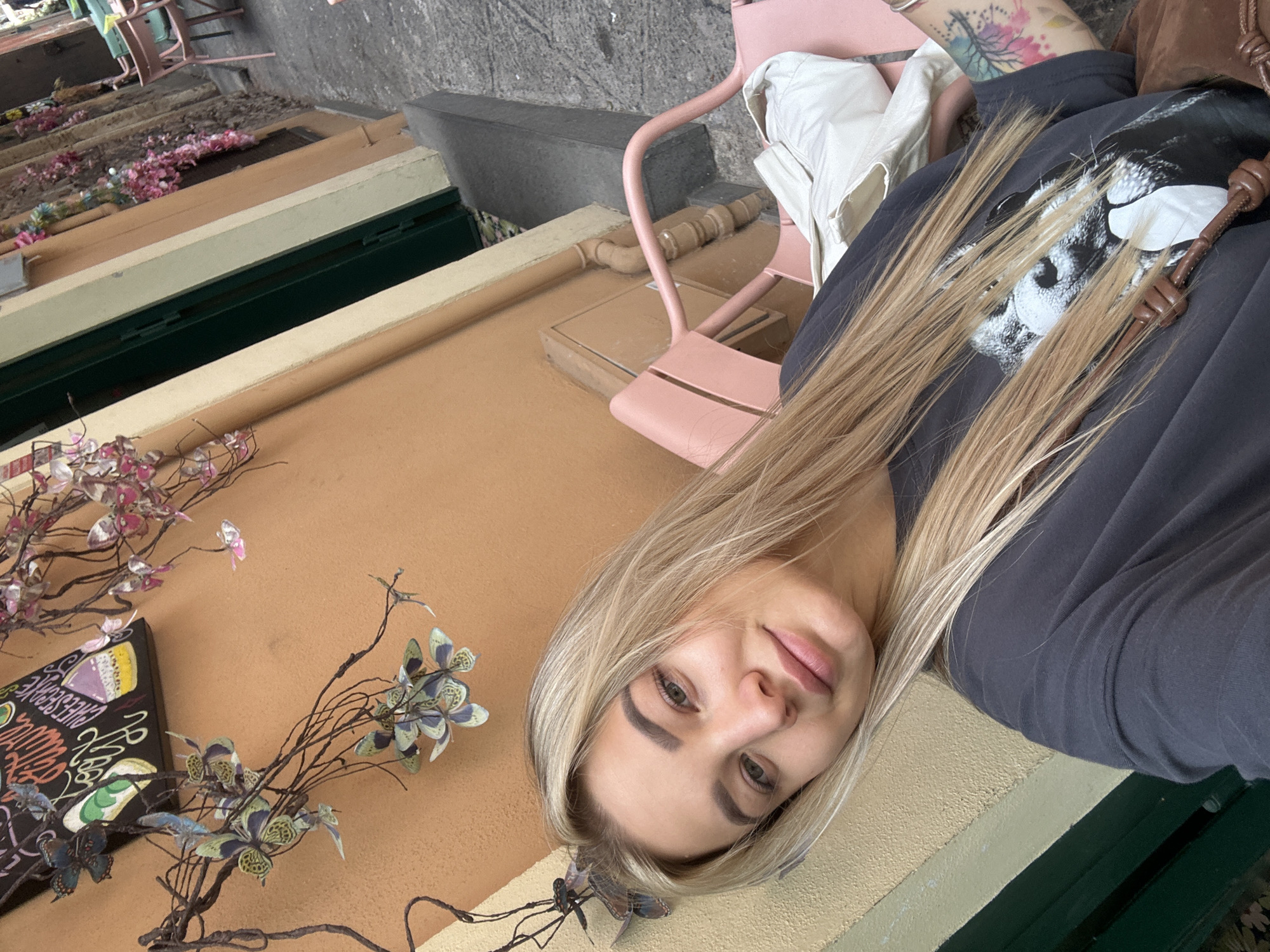 A medium-angle shot depicts a blonde woman taking a selfie in what appears to be an outdoor setting. She is wearing a dark-colored t-shirt with a graphic design. The background consists of a concrete surface and layered structures painted in shades of brown, white, and green. Pink floral decorations are also visible. To the right of the woman, there is a pink chair. Based on the overall characteristics, the location is likely in Catania, Italy.