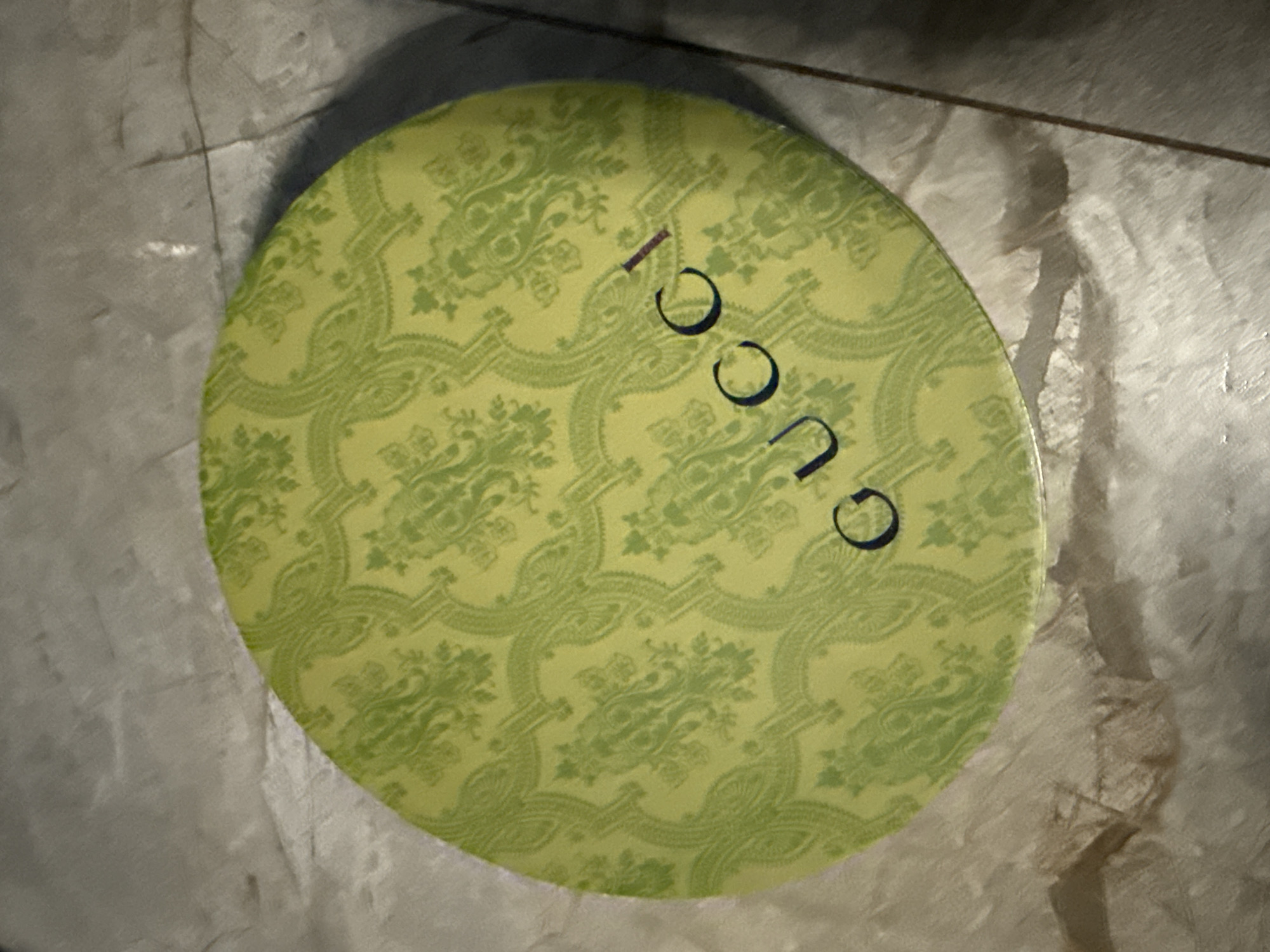The scene, located in the city of Berezyna, Ukraine, depicts a round, lime-green object with a darker green damask-style repeating pattern resting on a light-colored, textured surface. The word "GUCCI" is printed in dark blue/purple capital letters across the lower right portion of the object. The object appears to be a lid or the top surface of a shallow container. The background surface consists of large, light grey or off-white tiles or concrete slabs with visible seams and a slightly uneven texture. A subtle shadow is cast along the lower right perimeter of the round object, indicating illumination from above and to the left. No people, actions, or interactions are visible in the image.