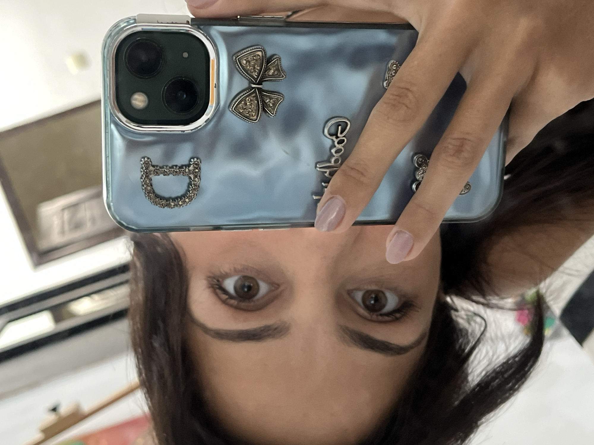 A woman is taking a selfie in an unknown location, possibly in Unknown, Indonesia. She holds a blue, bedazzled phone above her face, partially obscuring her forehead. The phone has three visible camera lenses. The phone case features a bedazzled butterfly, the word "Good", and other decorative elements. The woman has dark brown hair, brown eyes, and dark eyebrows. She has light-colored nail polish on her fingers. A picture frame is visible in the background, out of focus. The environment is otherwise indistinct.