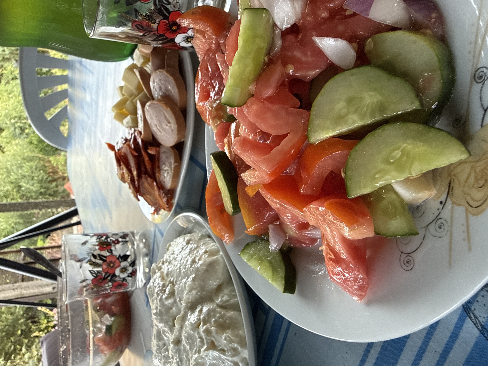 An outdoor table setting features a meal in Ekzarh Antimovo, Bulgaria. A white plate in the foreground holds a salad composed of chopped tomatoes, cucumbers, and red onion. To the left, a white bowl contains a creamy dip. In the background, another plate holds slices of sausage, bacon, and cubed cheese. Two decorative glasses with floral patterns are visible, one with a spoon inside. A green pitcher sits behind the glasses. The table is covered with a blue and white striped tablecloth. The background shows greenery and the partial view of a white chair, suggesting a natural or garden setting. There are no people in the image.