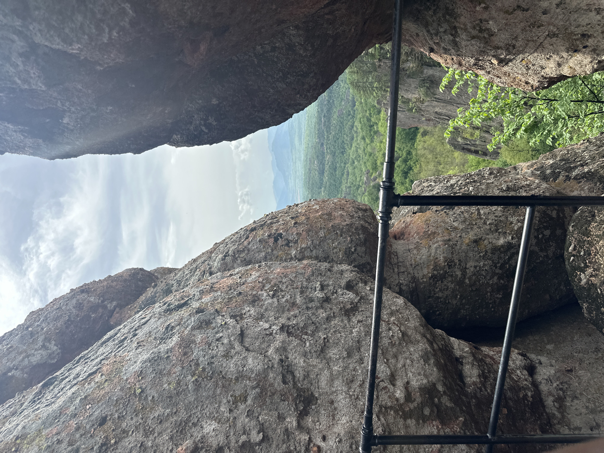 The image shows a view through a narrow opening between massive rock formations in Belogradchik, Bulgaria. The foreground is dominated by the rough, weathered surfaces of the rocks, and a section of a black metal pipe ladder running diagonally across the frame. Through the opening, a green landscape stretches out, consisting of forests and fields, with mountains faintly visible in the distance under a cloudy sky.