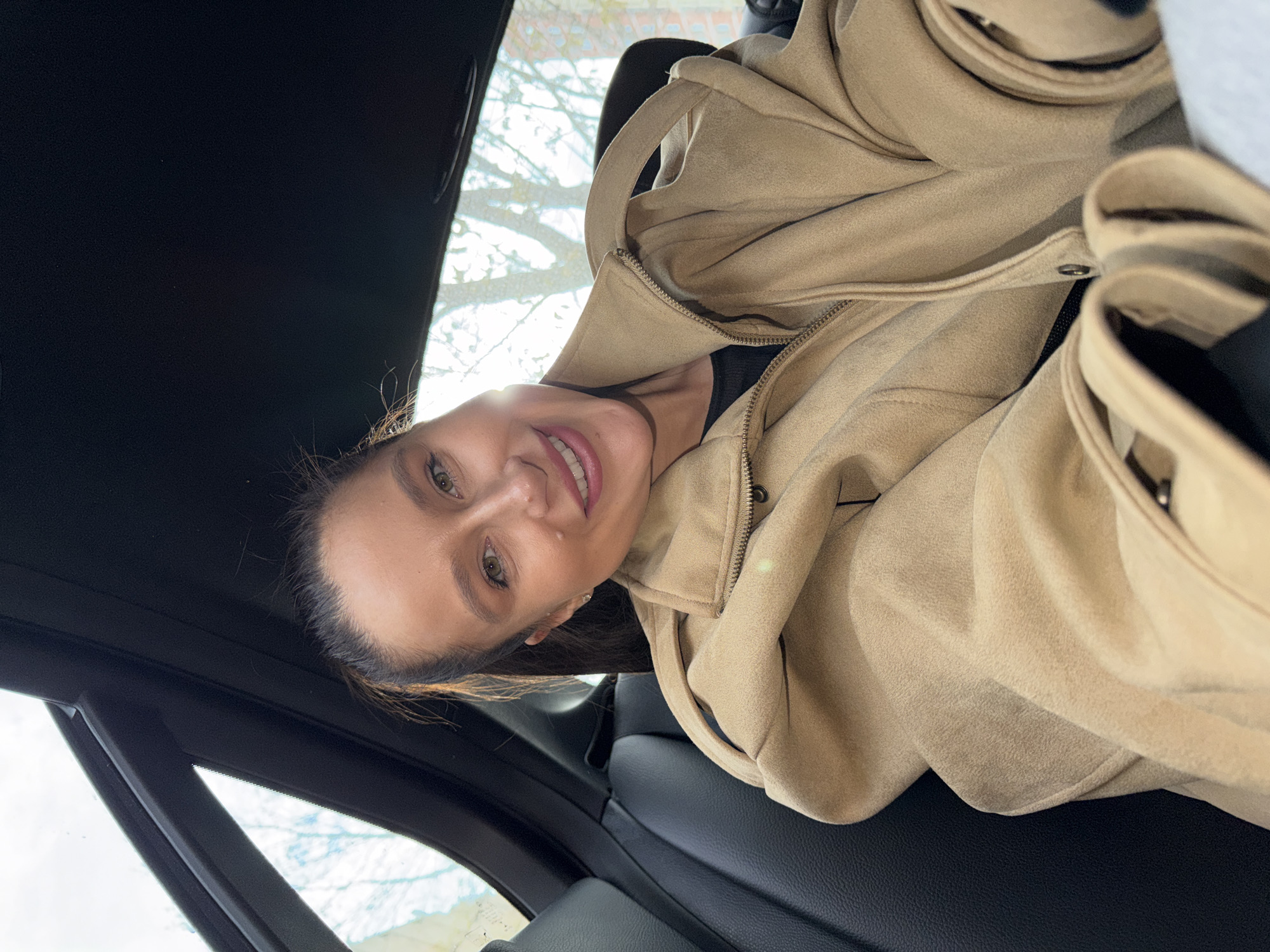 The image displays a smiling woman inside a car, looking directly at the camera. She has dark hair pulled back and striking green or hazel eyes. She is wearing a light beige or tan jacket with a zipper, over a dark-colored top. The interior of the car is visible, featuring a dark headliner and black leather seats. Through the car windows, a bright, possibly sunny, daytime sky is visible, along with what appear to be bare tree branches, suggesting a cooler season like winter or early spring. The woman's expression is pleasant and happy. The image is rotated 90 degrees clockwise. The scene takes place in Khimki, Russia. There is no visible text in the image.