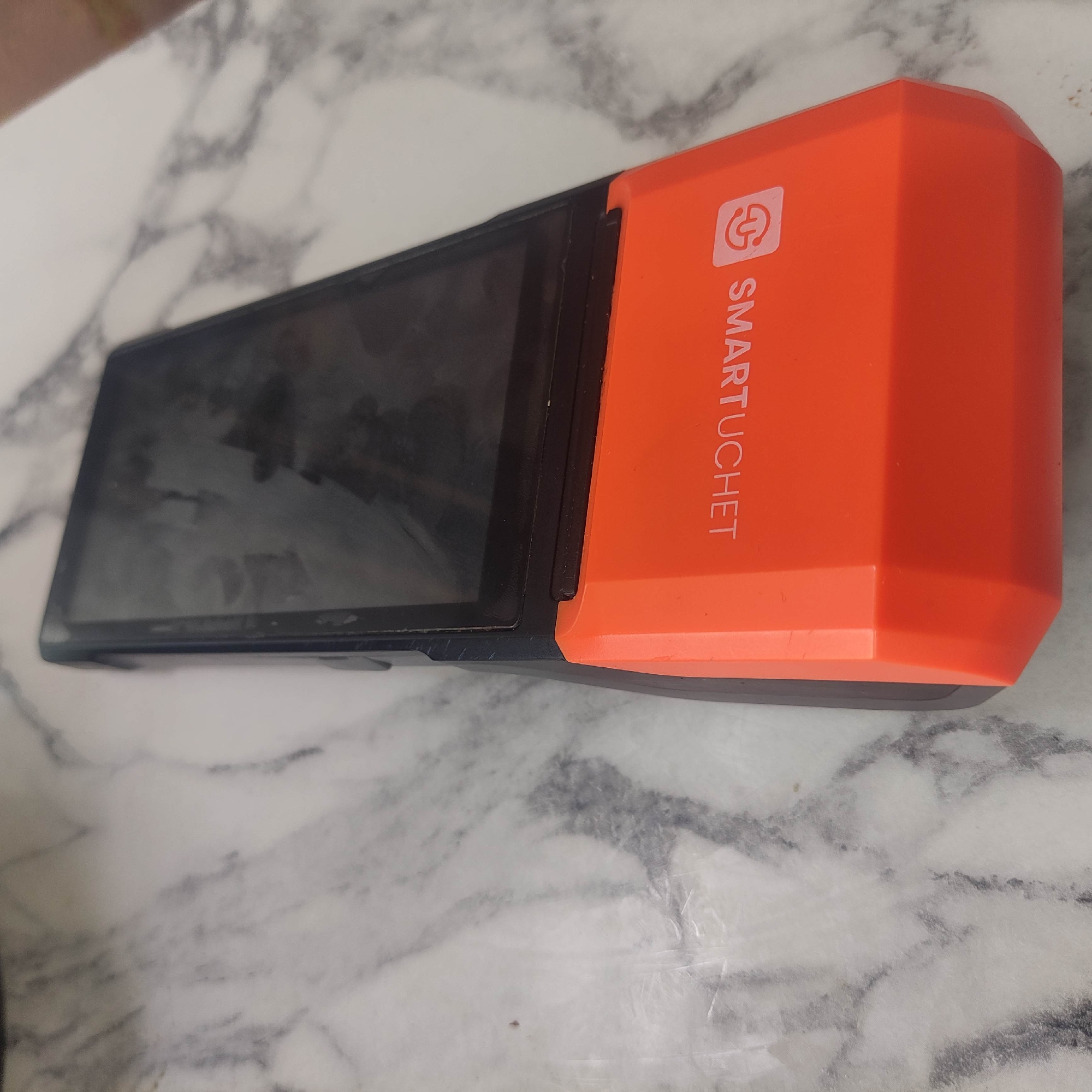 The image shows a close-up, angled shot of an orange and black point-of-sale (POS) device resting on a marble-patterned surface. The device has a black screen at the top and a bright orange lower section with white text that reads "SMARTUCHET" and a logo. The surface appears to be a countertop or table. The lighting is somewhat dim, casting soft shadows. There are no people visible in the image. The background shows the marbled texture of the surface. The location is identified as Razzakov, Kyrgyzstan.
