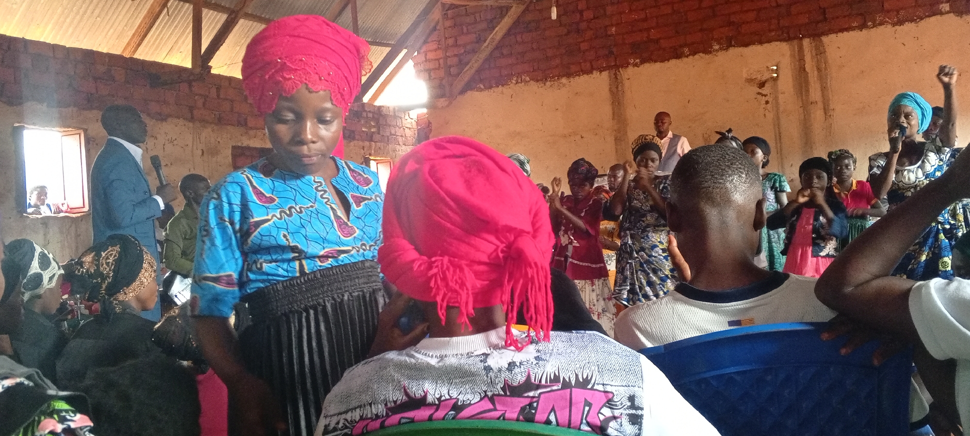 The image shows an indoor scene, possibly in a church or meeting hall in Ngalula, Democratic Republic of the Congo. The building appears to be constructed of brick with a metal roof. A man in a suit is standing and speaking into a microphone near a window. Several people are present, some seated and others standing. Many of the women are wearing colorful headwraps. Some individuals are raising their hands, possibly singing or praying. A woman in a blue patterned top and red headwrap stands in the foreground, while another individual with a red headwrap sits facing away from the camera.