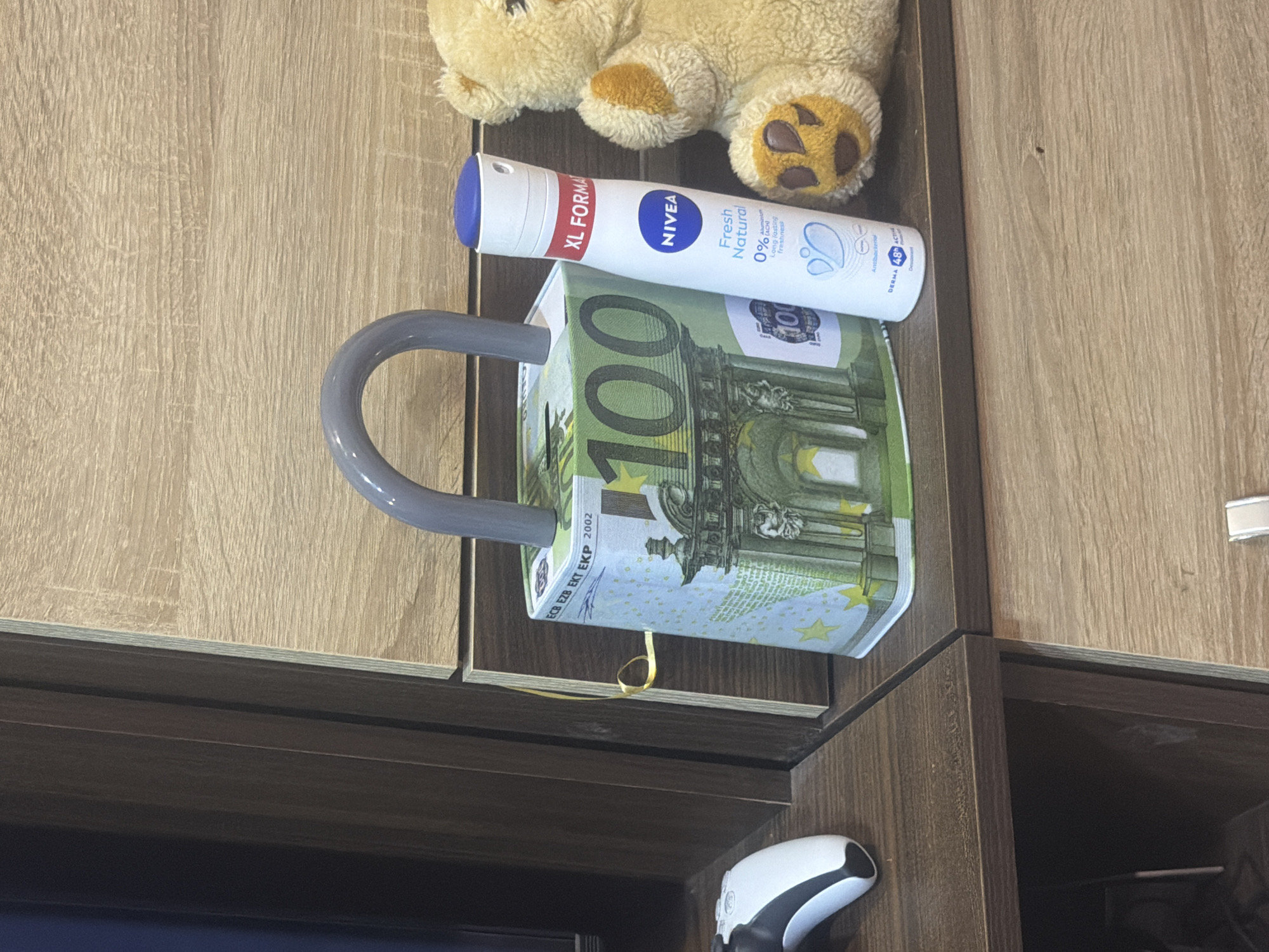 An indoor scene displays various objects arranged on and around light brown wooden furniture, possibly cabinets or shelves. The upper portion of a beige stuffed animal with brown paw pads is visible in the top left corner, resting on the wooden surface. Below it, positioned horizontally, is a white spray can, likely a deodorant, resting on top of a distinctive money box.
The money box is shaped like a mug with a grey handle and is covered with a design mimicking 100 Euro banknotes, featuring the numerical value "100" prominently. A thin yellow ribbon hangs from the bottom edge of this money box. Below the money box, partially visible at the bottom right, is a white and black object that resembles a modern gaming controller. The background consists of vertically paneled wooden surfaces of varying shades, suggesting different parts of the furniture.
Visible text on the spray can includes "XL FORMA", "NIVEA", "Fresh Natural", "0%", "Long lasting freshness", "Antibacterial", and "DERMA ADAC". Text visible on the money box, mimicking a 100 Euro banknote, includes "ECB EZB EKT EKP 2002" and the number "100".