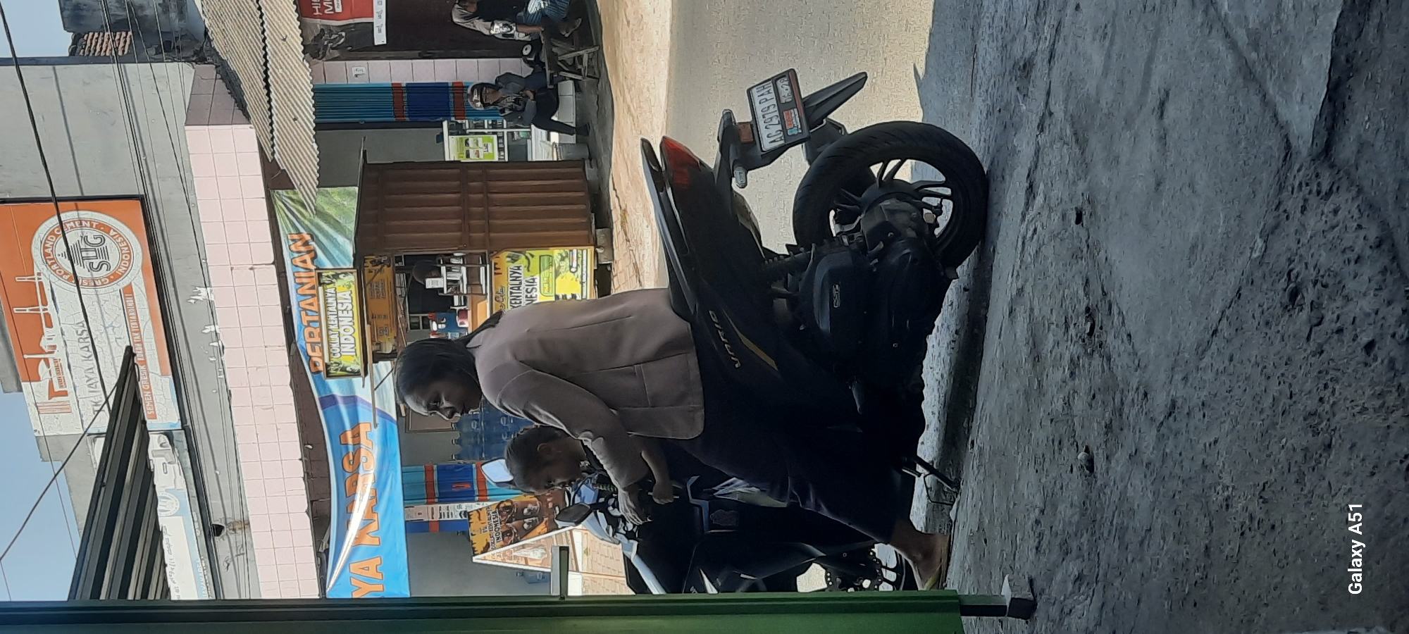 This is a medium shot, eye-level photograph taken outdoors in an urban environment, likely in Selopuro, Indonesia. The foreground features the rear of a black motorcycle, with a license plate that reads "AG 2979 PAH". On the motorcycle, a woman wearing a beige jacket and dark pants is seated behind the handlebars, with a young child in a dark outfit positioned in front of her. The woman's head is turned to her right, and she appears to be looking off-camera. The child is looking towards the left.
The background shows a building with a pastel pink facade, a corrugated metal roof, and several awnings. A banner with Indonesian text, including "PERTANIAN" and "INDONESIA", is visible. To the right of the building, a dark brown kiosk-like structure with glass panels is present, and a person wearing a helmet and dark clothing is seated on a low step. Further to the right, two women are sitting on a wooden bench, one wearing a hijab. The ground is paved with a rough, grayish surface, and the sunlight casts shadows across the scene. The overall impression is of a busy street or market area.