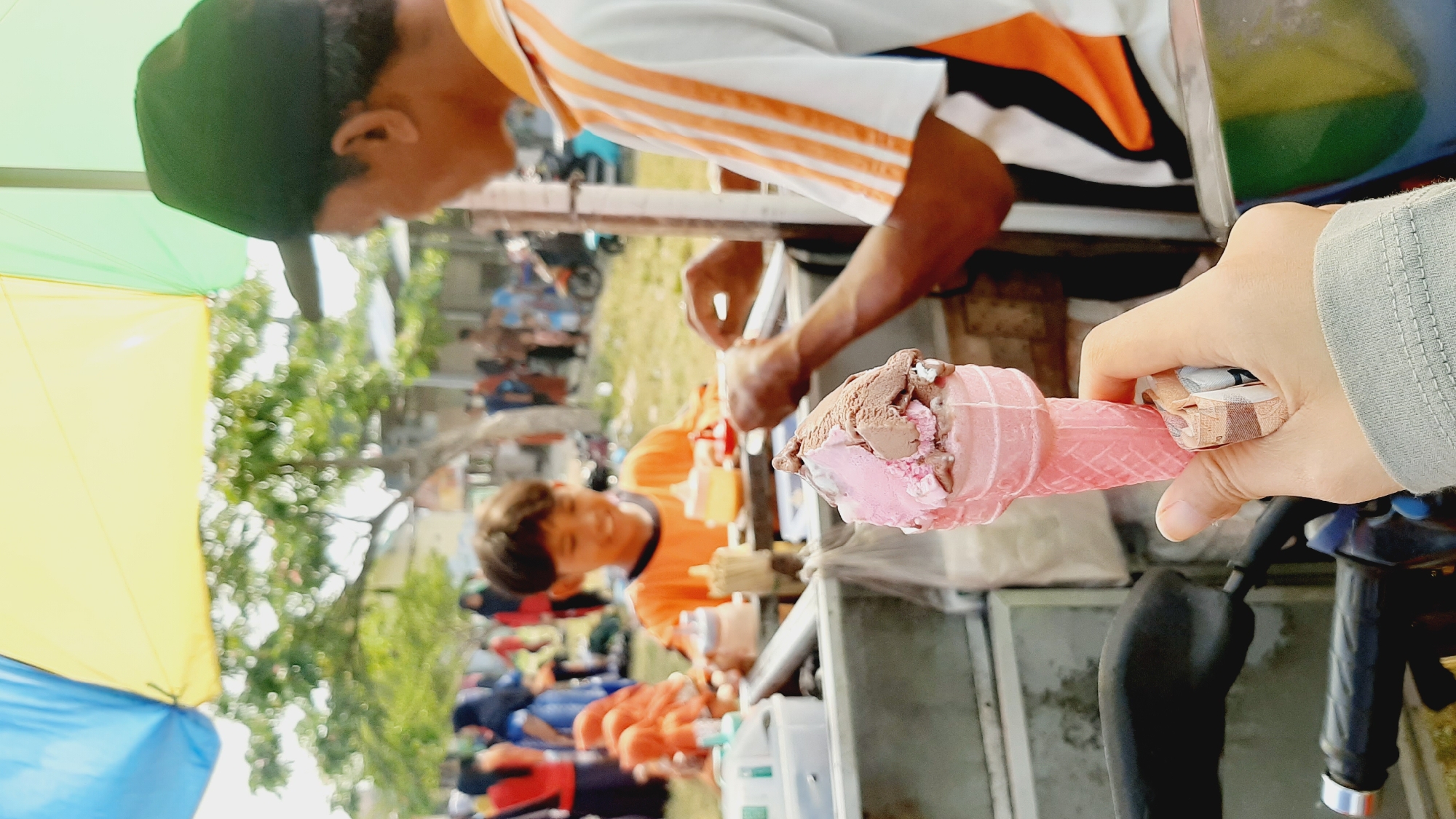 In Sumberjo, Indonesia, a person's hand holds a pink ice cream cone with two scoops of ice cream, one pink and one chocolate brown, and some folded currency. The hand is wearing a grey long-sleeved shirt. In the background, an ice cream vendor, wearing an orange and white striped shirt and a dark green baseball cap, is preparing or serving ice cream. A young boy in an orange shirt is smiling and appears to be a customer. Other people and a parked motorbike are visible in the blurred background, suggesting a public outdoor setting, possibly a market or street fair, with a multi-colored umbrella providing shade.