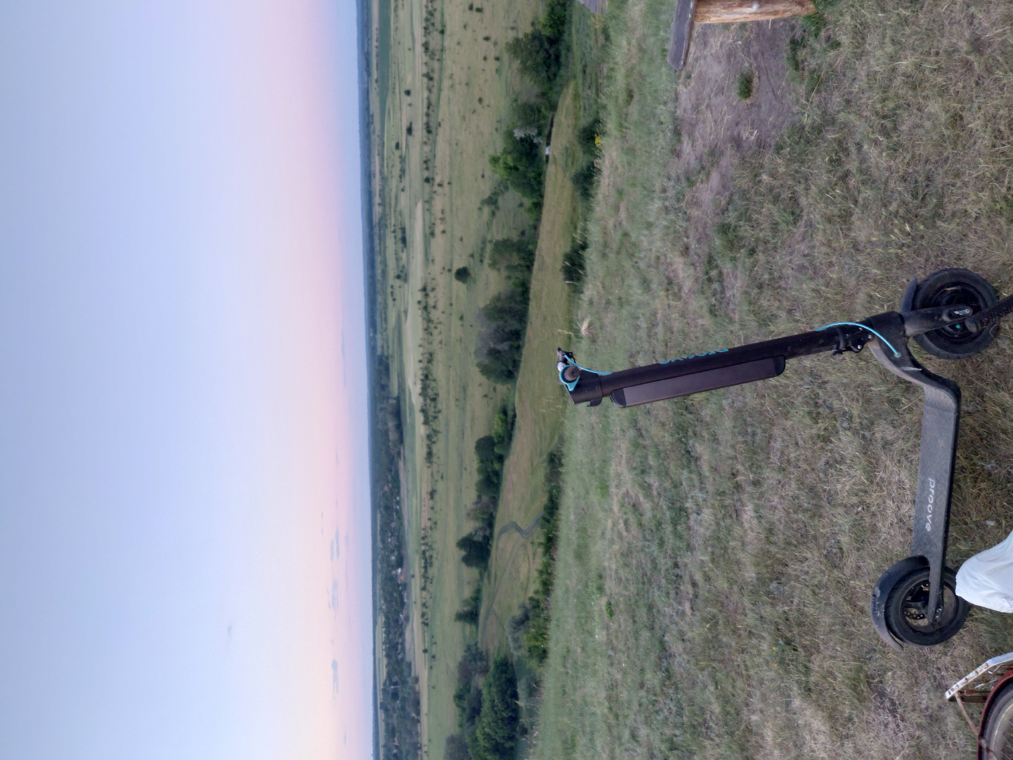 An electric scooter is parked on a grassy hilltop overlooking a rural landscape at dusk. The sky is a gradient of pale blue to pink, with distant clouds. The scooter is black with blue accents and has the word "PROOVE" printed on its deck. To the right, a wooden bench made of a log and plank is visible. The landscape below is a patchwork of fields and trees, with a winding path visible in the middle ground. The scene appears to be in or near Ustyvytsia, Ukraine, given the context.