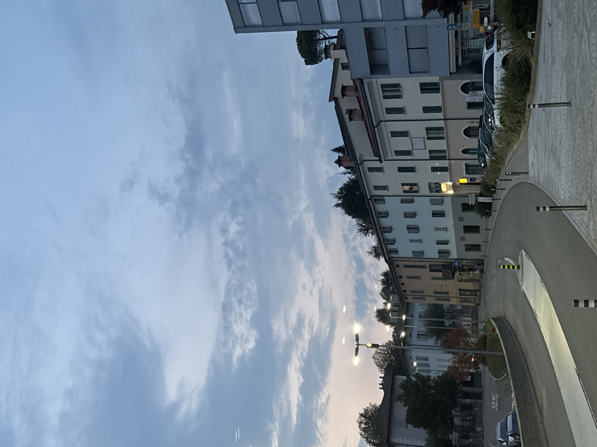 The image shows a street scene in the city of Balerna, Switzerland, during twilight. A row of multi-story buildings with colorful facades lines the right side of a curved road. The buildings have windows with green shutters and some have balconies. Streetlights are visible, some illuminated. Several cars are parked along the street, and a few people are walking on the sidewalk. The road has painted lane markings and bollards. The sky is cloudy and the light suggests it is either early morning or late evening. Trees and other vegetation are present, bordering the buildings and the road. The architecture suggests a European urban setting.