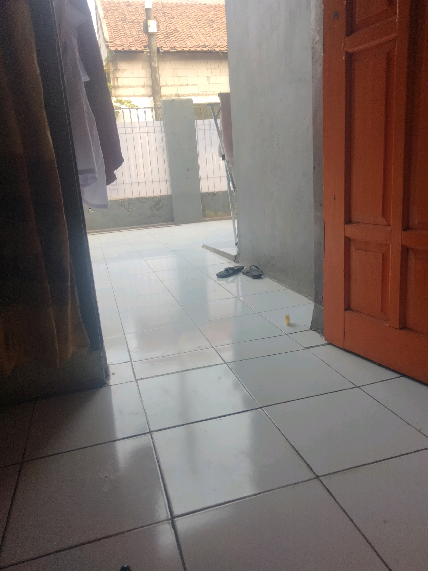 This image depicts an outdoor view from a doorway in Suka Sari, Indonesia. The scene shows a tiled floor leading outwards, with a portion of an orange door visible on the right. To the left, a brown curtain hangs, partially obscuring a white garment and a dark red garment. In the background, a white fence with vertical slats is visible, with a building and a reddish-brown tiled roof beyond it. A light pole with a white box is also present. On the tiled floor in the middle ground, a pair of black flip-flops and a small yellow object are visible. The lighting suggests it is daytime.
