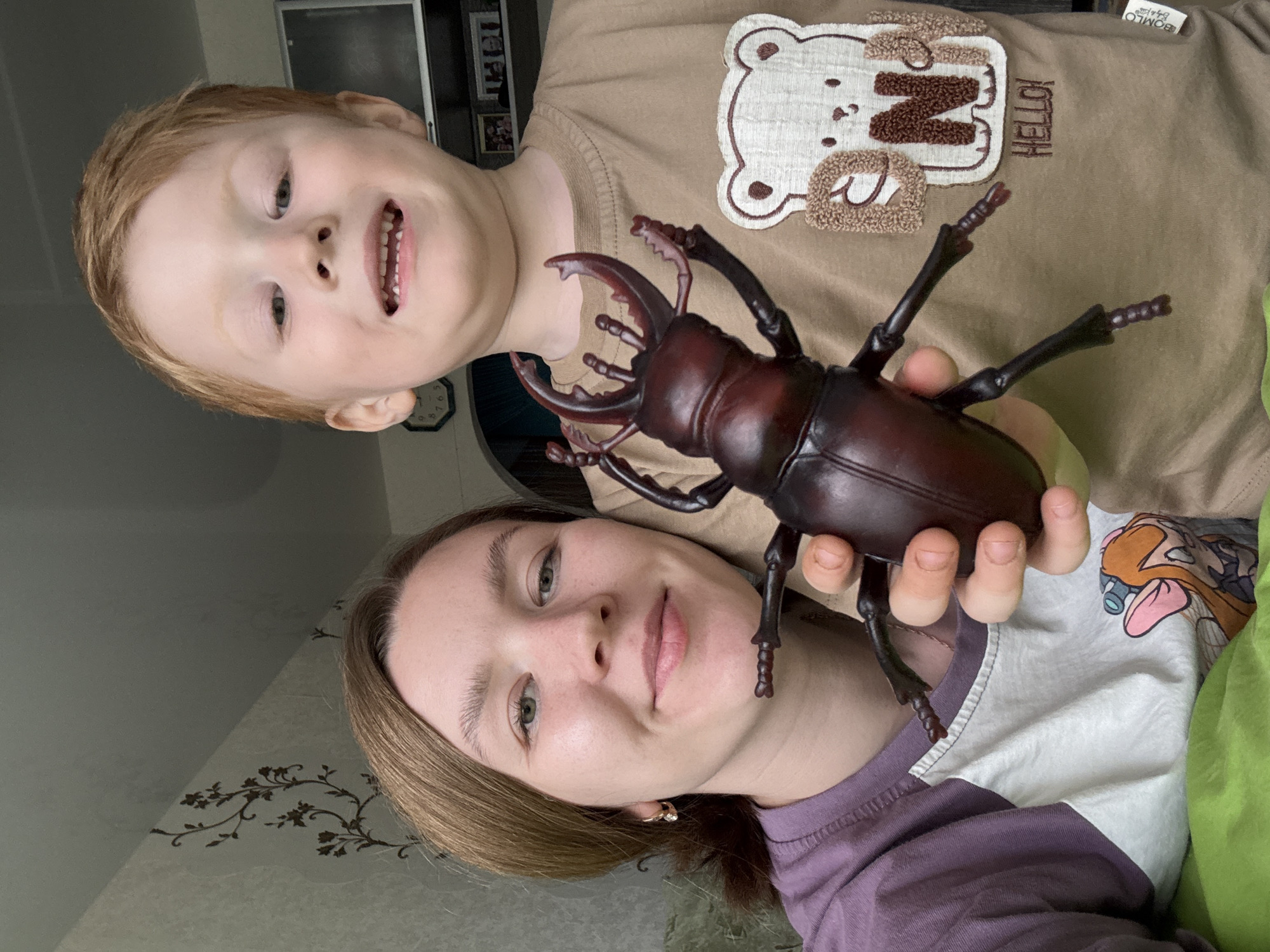 A woman holds a large, dark brown beetle toy. A young boy with red hair stands behind her and to the left, smiling. The woman is wearing a purple shirt and the boy is wearing a tan shirt with a bear design. The photo appears to be taken indoors, near a wall with a floral design. The location is Chaikovsky, Russia.