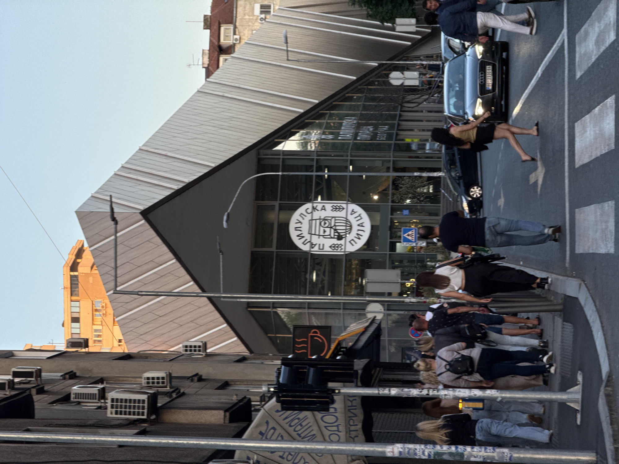The image shows a street scene in the Palilula Urban Municipality, Serbia, likely during the daytime given the lighting. A modern building with a distinctive angular, metallic facade dominates the right side of the frame. The building features large glass windows. In front of this building, a circular logo with stylized imagery and Cyrillic text is visible. To the left, an older building with multiple air conditioning units attached to its exterior is partially visible.
Several people are walking on the sidewalk and crossing the street. One group of individuals is walking away from the camera on the left sidewalk. Another group is crossing a pedestrian crossing in the foreground, with one woman in a light-colored top and dark pants walking across. A dark-colored Audi sedan is stopped at the pedestrian crossing, with its headlights on. Other cars are visible in the background.
A traffic light with multiple signals is mounted on a pole on the left side of the image, and a sign with an illuminated coffee cup graphic is attached below it. Streetlights are also visible. The sky is clear and blue. The overall impression is an urban environment with a mix of modern and older architecture.