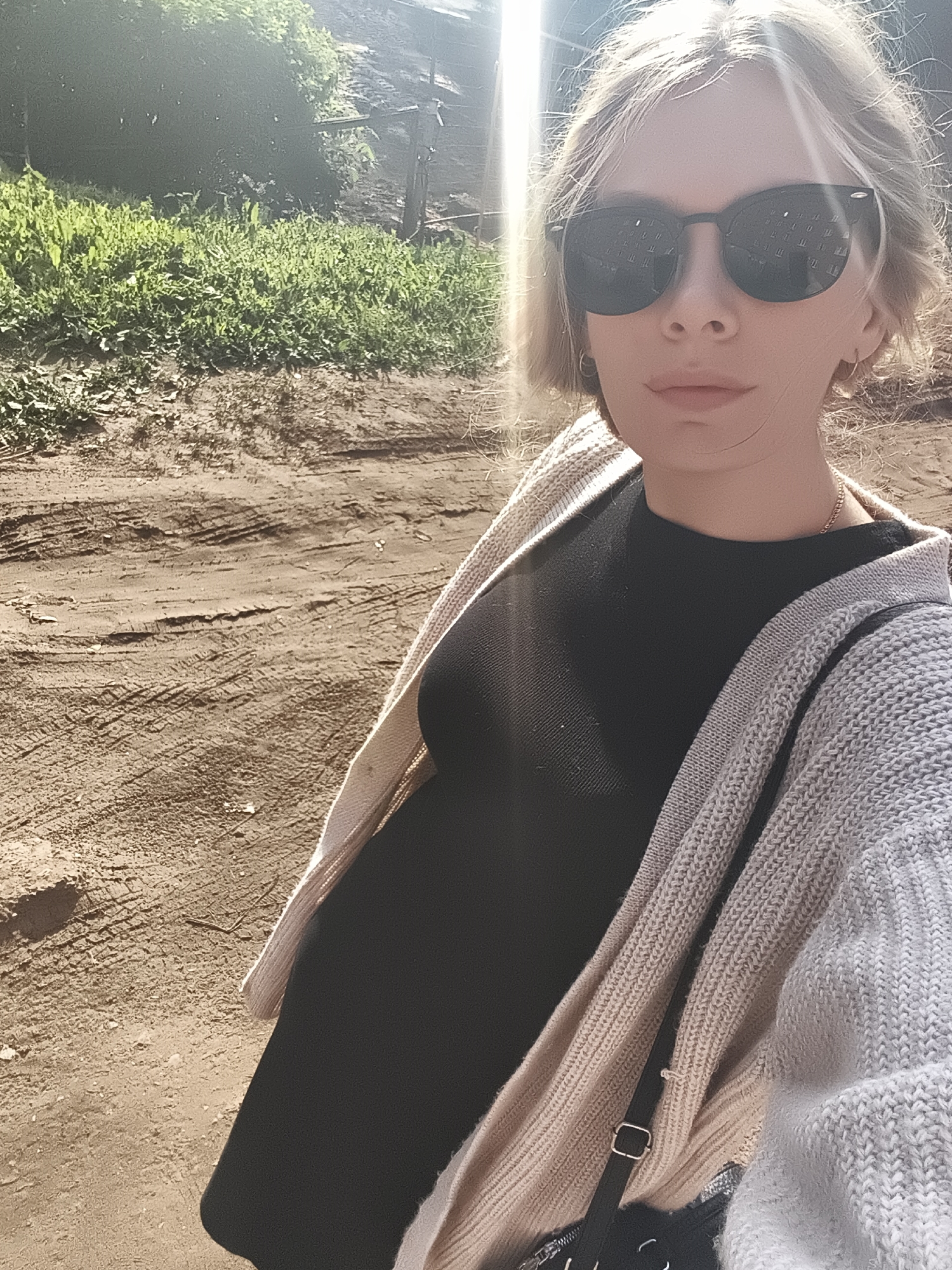 The image shows a fair-skinned person wearing sunglasses, a black dress, and a beige cardigan, standing on a dirt road. The background features green vegetation and a wooden structure. The location appears to be outdoors, possibly in a rural or semi-rural area. There is no information in the image to conclude the location is in the city of Izhevsk, Russia.