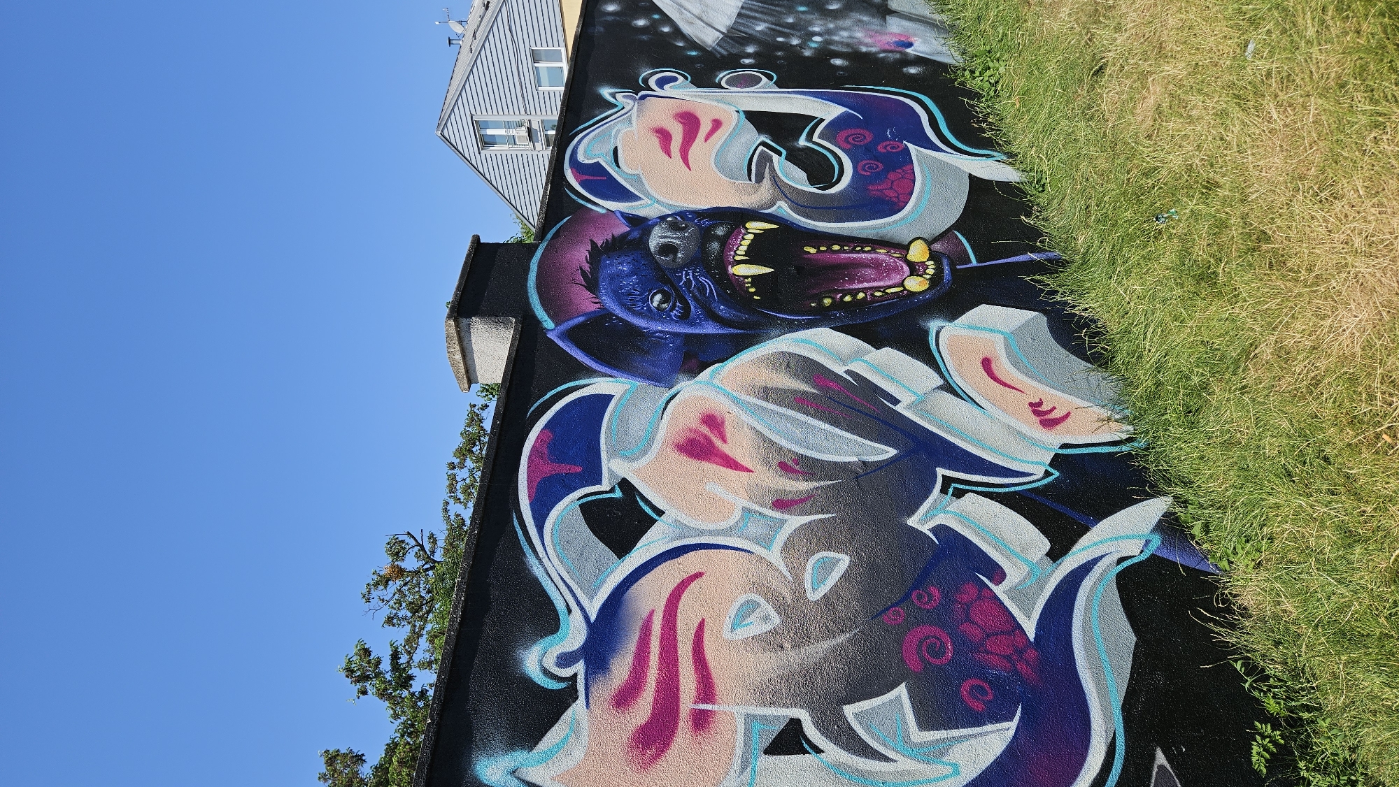 The image displays graffiti on a dark, textured wall in Prešov, Slovakia. The graffiti features a stylized depiction of a blue creature resembling a hyena with its mouth open, revealing sharp teeth and a purple interior. The creature's eyes are wide and yellow, and its face is rendered with detail. To the left of the creature, abstract graffiti lettering in shades of purple, pink, blue, and light gray is visible, with intricate swirls and shapes. The wall is bordered by a strip of green grass in the foreground, some of which appears dry and yellowed. Behind the wall, trees with green leaves and some orange berries are visible against a clear, bright blue sky. A portion of a building with light blue siding and white trim, featuring windows, is present in the upper right corner of the frame. A grey concrete chimney or ventilation structure is also visible on top of the wall. The lighting suggests it is a sunny day.