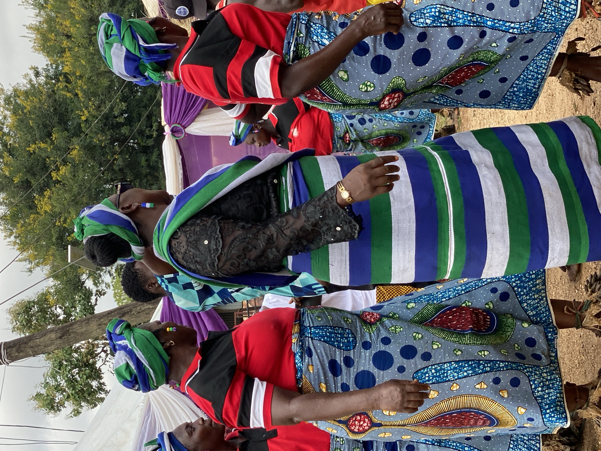 In an outdoor setting in Maigizo, Nigeria, multiple individuals are present. All are wearing traditional African attire. The outfits are colorful, with distinct patterns on head wraps and other clothing. One woman wears a lace top and has a gold watch. The setting includes a tent with purple and white decorations, and trees are visible in the background.