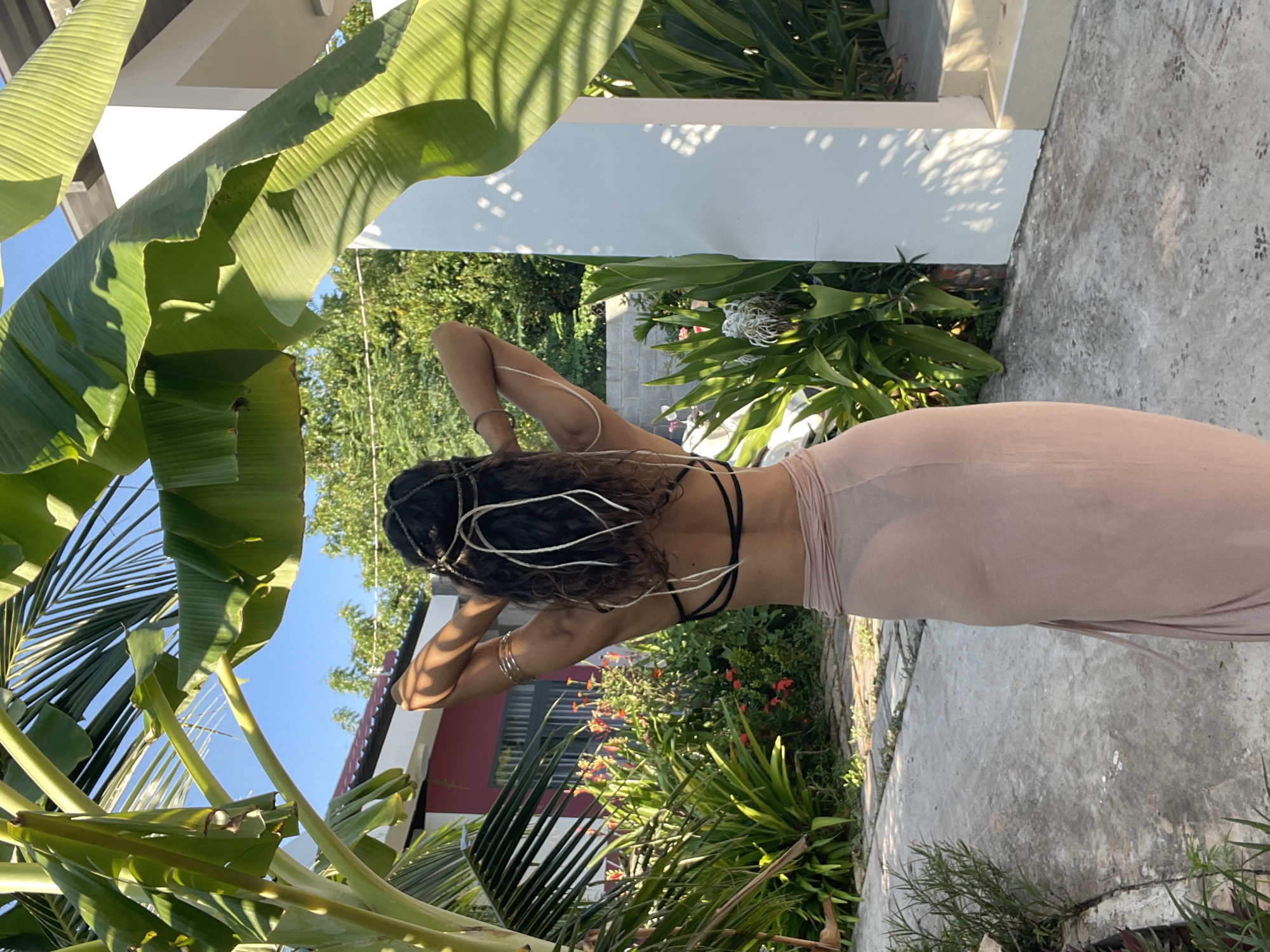 A person is standing outdoors in what appears to be a tropical setting, possibly in Mũi Né, Vietnam. The person is viewed from behind, wearing a black bikini top and a long, light pink skirt that is semi-sheer. They have their arms raised behind their head, and their hair is dark and curly, adorned with thin metallic strings. The background features lush green foliage, including large banana leaves, with a house visible in the distance. The ground is a paved pathway.