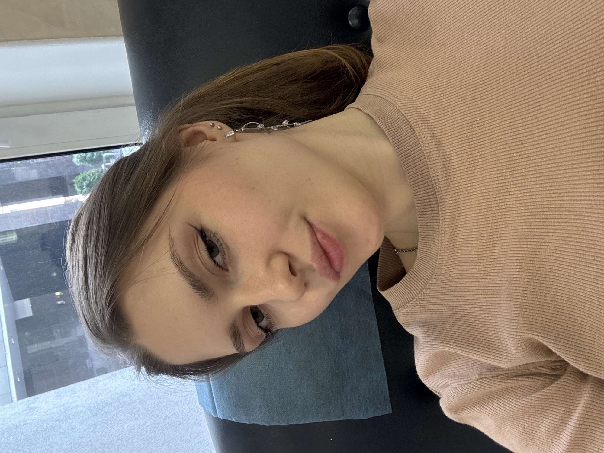A young woman is depicted in a close-up, angled portrait, appearing to lie down or lean back. Her head is tilted, and she looks directly towards the camera with a calm expression and a slight, gentle smile. She has light brown hair, which is styled to reveal her right ear.
Her ear is adorned with multiple silver-toned piercings, including several small studs along the helix and conch, and a distinctive, elongated decorative earring extending upwards from the lobe. This earring features several small, sparkling elements, possibly crystals or rhinestones. She wears a light brown or beige ribbed long-sleeved top or sweater, and a thin silver chain necklace is visible around her neck. Her makeup is subtle, with winged eyeliner, mascara, and light pink lip color.
The setting is indoors, with natural light suggesting it is daytime. Behind her, a large window reveals an urban landscape with buildings and some green foliage. Her head rests on a dark surface, which is partially covered by a blue disposable sheet, often used in professional settings like clinics, beauty salons, or piercing studios. No text is visible in the image. The scene is set in Saint Petersburg, Russia.