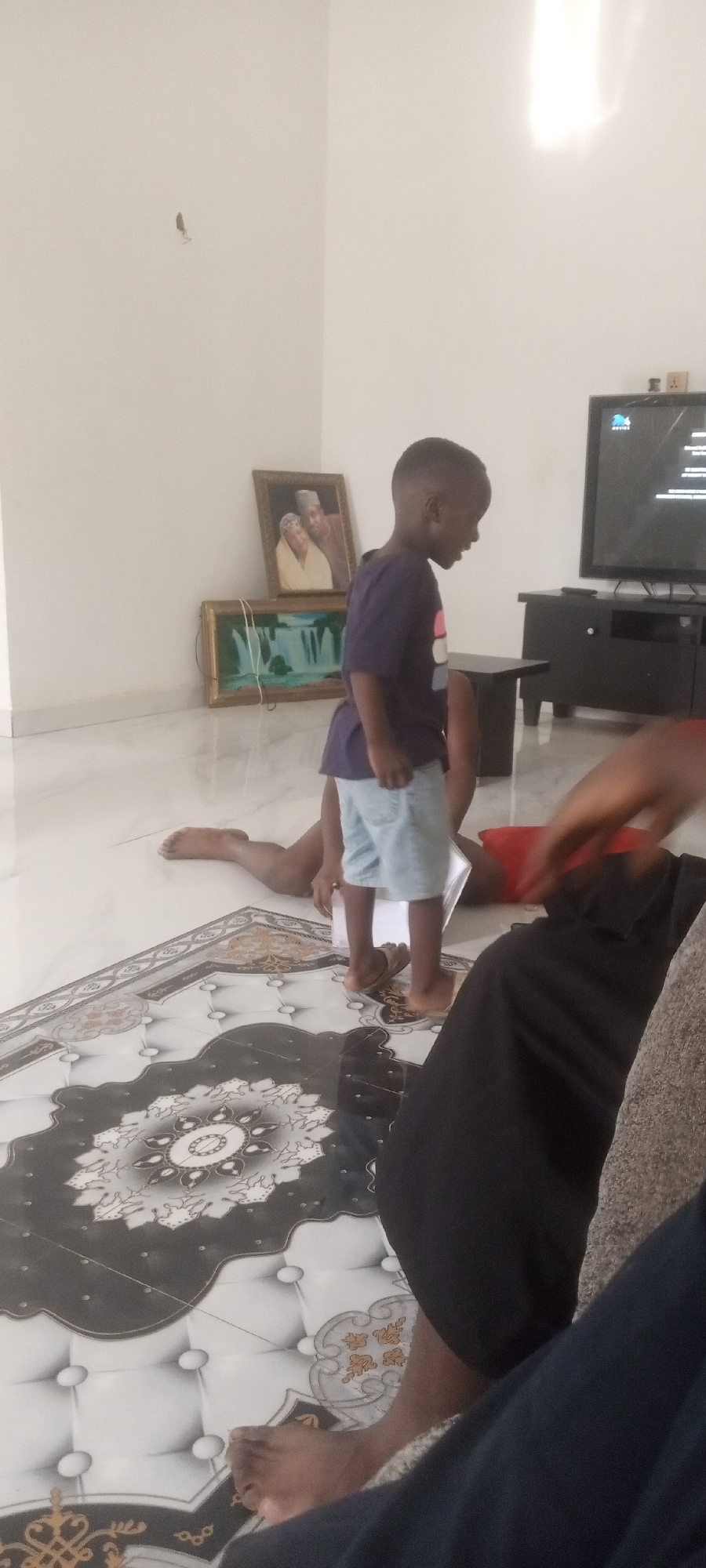 The image depicts an interior space with a child standing in the foreground, positioned near a person lying on the floor. The child is wearing a dark t-shirt and light-colored shorts, and they are barefoot. The individual on the floor is partially visible, with only their legs and torso discernible. The floor is tiled with a decorative pattern. In the background, there is a television set on a dark-colored stand, along with framed pictures leaning against the wall. The environment appears to be a residential living room. No specific location is indicated beyond "Unknown, Unknown". The presence of people in the image indicates activity, but no specific actions or interactions can be determined beyond the child's standing posture and the adult's reclining position.