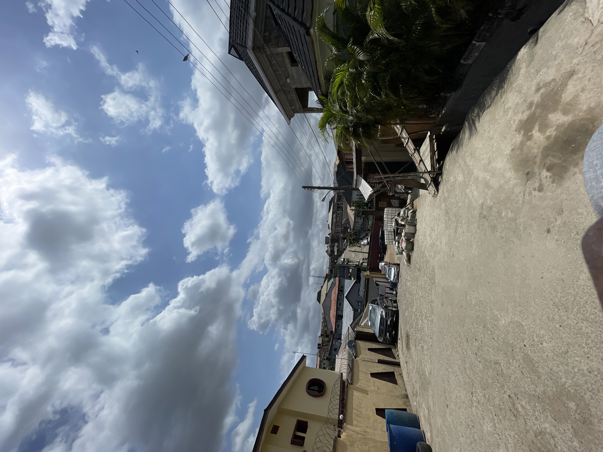 The image shows a street in Isheri, Nigeria, lined with buildings. The street is paved and appears to be moderately wide. Several parked cars are visible. The sky is partially cloudy with patches of blue sky visible. Power lines run above the street. Some buildings are under construction. There are no people visible in the image. The overall impression is a typical residential or mixed-use street scene in a developing urban environment.