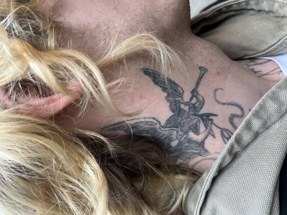 A close-up shows a person with blonde, wavy hair and fair skin. The focus is on their neck and upper chest, revealing an angel tattoo playing a trumpet while holding an olive branch. The tattoo is detailed and shaded in black ink. The person is wearing a beige garment, possibly a jacket or shirt. The composition emphasizes the tattoo's placement and the person's features.
