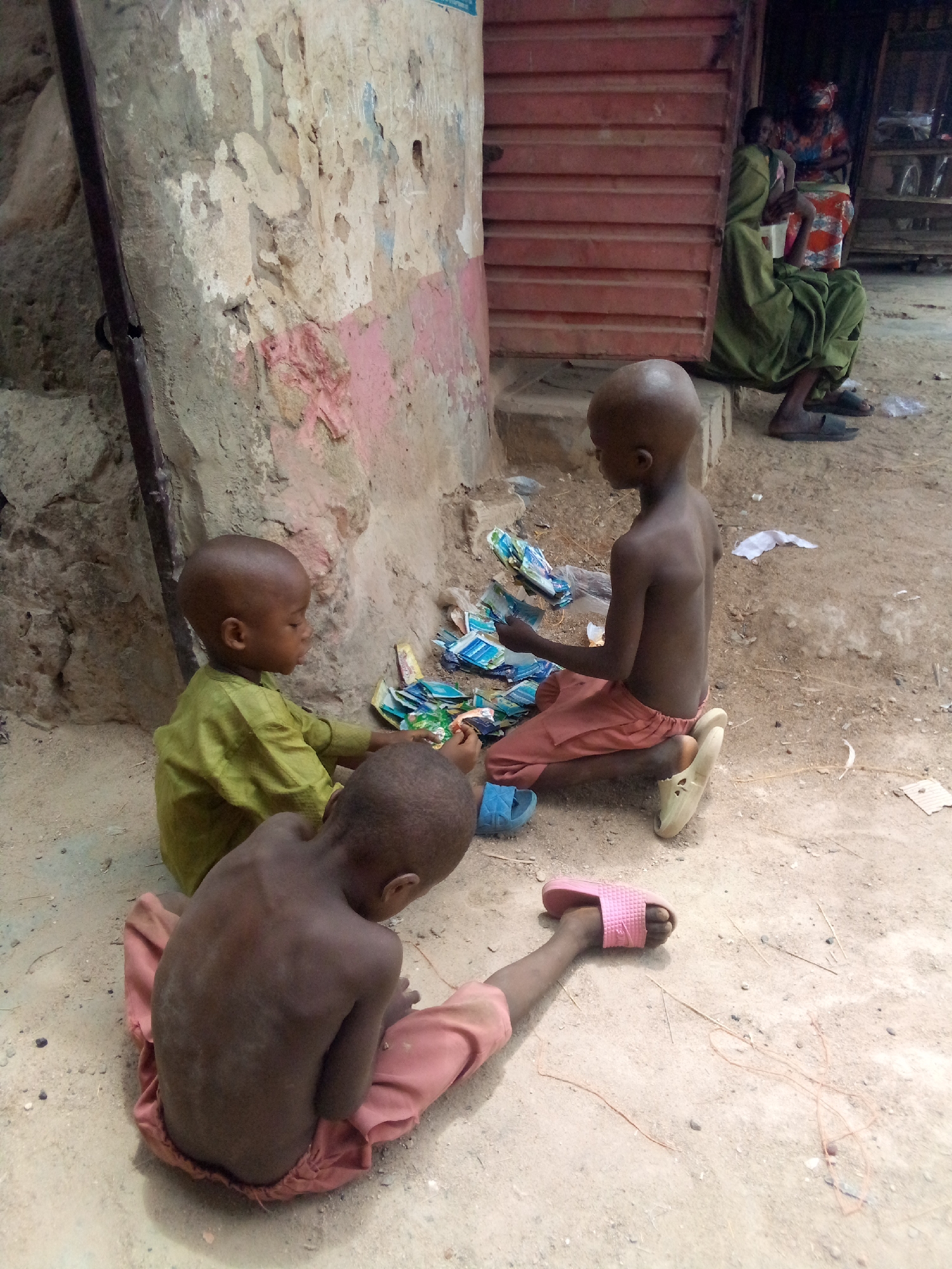 Three children are seated on the ground outside a building in Minna, Nigeria. The children are thin and appear to be playing with small, colorful packaged goods. One child wears a green shirt, another pink pants, and the third pink pants and a pink sandal. The building has a weathered wall with peeling paint. Behind the children, an adult sits outside a doorway.
