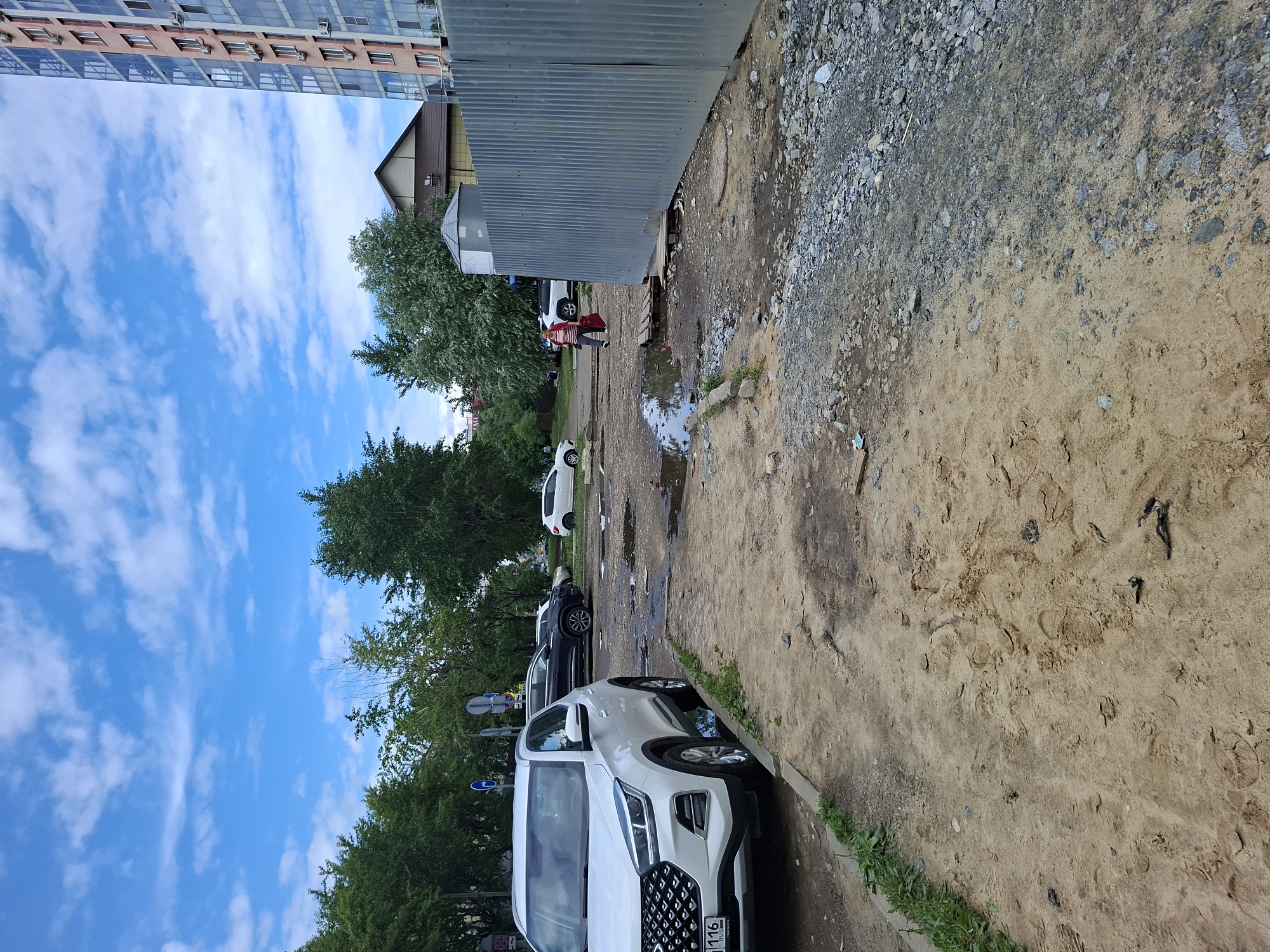 This media file depicts an outdoor scene on a cloudy day, likely in Kazan, Russia. The foreground is dominated by a sandy, unpaved area with patches of gravel, interspersed with puddles of water suggesting recent rain. A white SUV is parked along a curb on the right side of the frame. In the background, a corrugated metal fence separates the sandy area from a paved pathway where several cars are parked, including a white sedan and a dark-colored car. Further in the background, lush green trees line the street, and a multi-story building with a light-colored facade and numerous windows is visible. Another smaller building with a brown roof is also present. A person wearing a red top and dark pants is walking away from the camera near the fence, carrying a red bag. The overall impression is a slightly neglected urban environment, possibly a residential courtyard or parking area. There is some visible text on the license plate of the white SUV, which appears to read "116".