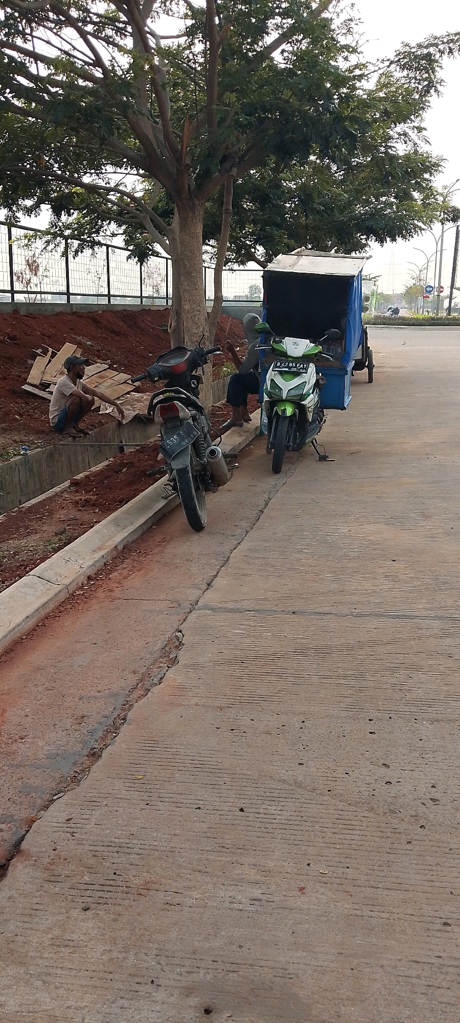 The image depicts an outdoor scene in Satriamekar, Indonesia, featuring two men, two motorcycles, and a trailer attached to one of the motorcycles. One man is crouching on the left, near a pile of reddish-brown dirt and some wooden planks. He is wearing a light-colored shirt and a dark cap. A dark-colored motorcycle is parked to his right, with its rear wheel facing the camera.
To the right of the dark motorcycle, a person wearing a grey hoodie and dark pants is sitting. In front of them, a green and white motorcycle is parked. This motorcycle is attached to a blue trailer, which appears to be a makeshift cargo carrier. The trailer has a metal frame and a blue tarp covering its contents.
The scene is set on a concrete surface, likely a road or pathway, with a concrete curb and a drainage ditch running along the left side of the image. Behind the men and motorcycles, there is a tree with extensive branches and foliage, and a black metal fence. In the background, there are signs and streetlights, suggesting a public or commercial area within the city. The sky is overcast.