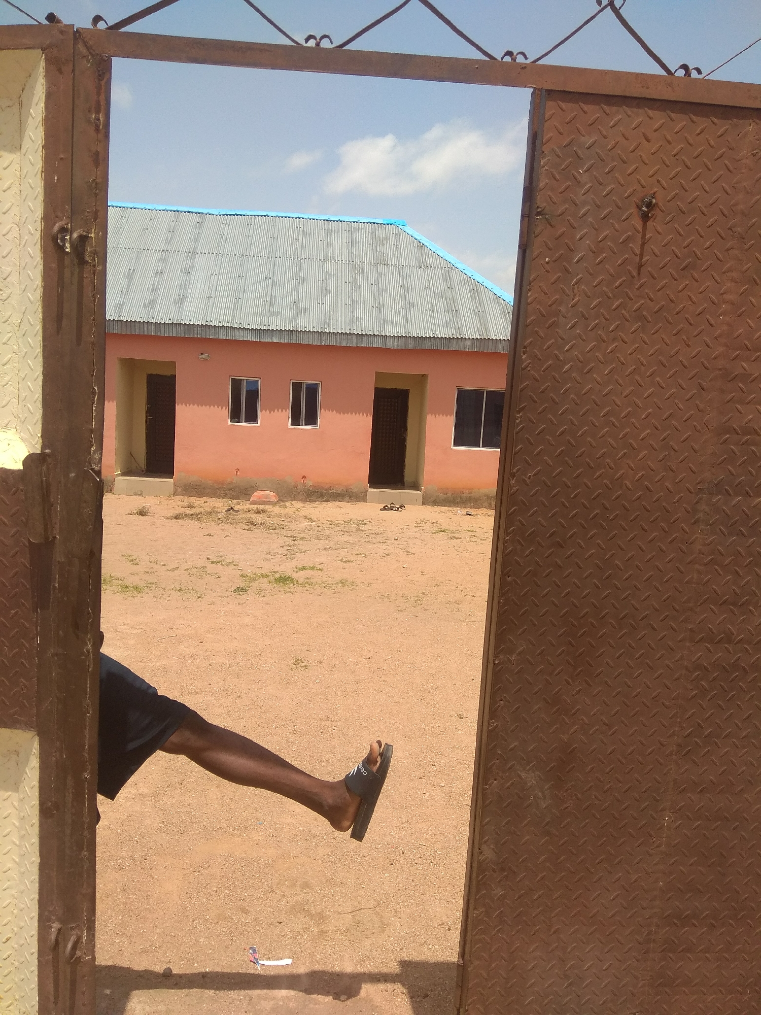 The image shows a person's leg, clad in dark shorts and a black flip-flop, extended outwards and resting on a dusty ground. The leg is positioned in the foreground, partially obscured by a rusty metal gate on the left. The gate is a prominent object in the frame, made of metal with a patterned texture. To the right of the gate, another section of a similar metal structure is visible.
Beyond the gate, a building with a pink facade and a corrugated metal roof with a blue trim is visible. The building has several windows and doors. The ground in front of the building is a sandy, dusty area with sparse patches of dry grass. The sky above is blue with some white clouds, suggesting it is daytime.
In the distance, on the ground in front of the building, a small, colorful object, possibly a toy airplane, is visible. The overall scene appears to be an outdoor setting, likely a residential area or a compound in Jalingo, Nigeria, given the provided location context. The lighting and shadows suggest it is likely midday or early afternoon. There is no visible text in the image. The image captures a casual moment, with the person's relaxed posture.